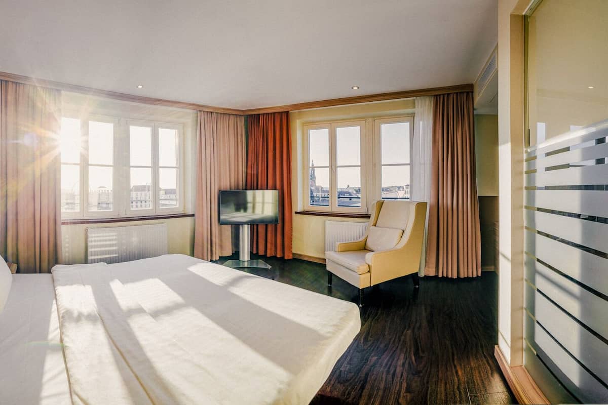 A hotel room with a large bed, a chair, a television, and a view of the city through the windows.