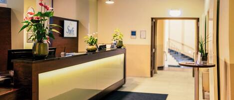 A hotel reception area with a reception desk, potted plants, and a staircase in the background.