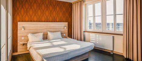 A hotel room with a large bed, wooden headboard, and a view of the city through large windows.