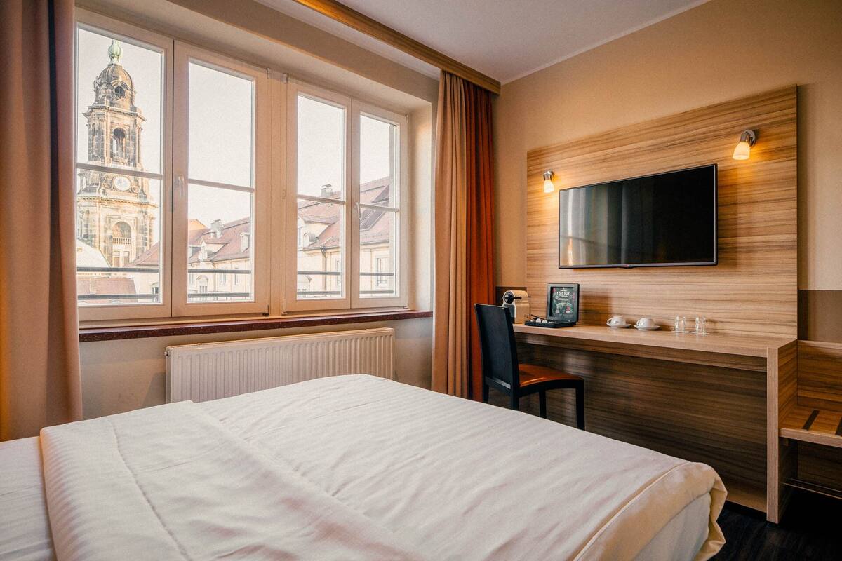 Hotellihuone, jossa on sänky, työpöytä tuolilla, seinälle asennettu televisio ja ikkuna, josta on näkymä rakennukseen.