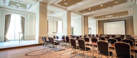 Une salle de conférence équipée de plusieurs tables et chaises, de grandes fenêtres et d’un écran de projection.