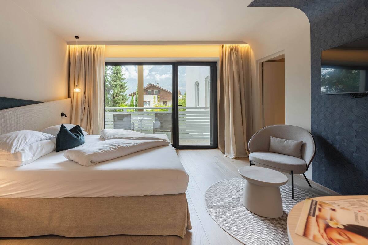 A modern hotel room with a large bed, a seating area with a round table, and a balcony with a view of the outside.