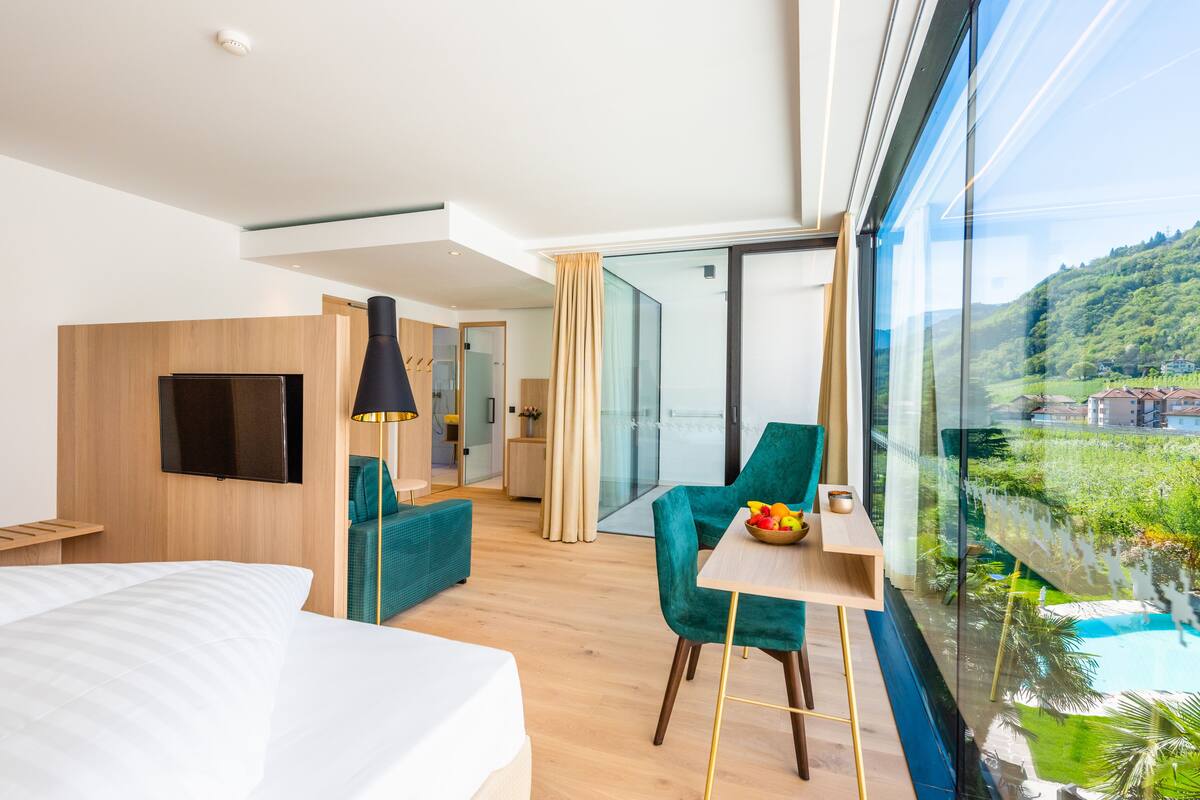A modern hotel room with a large bed, a TV, a dining area with a table and chairs, and a view of a lush landscape through large windows.