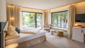 A modern hotel room with a large bed, a small seating area, a desk, and large windows offering a view of palm trees. - Hotel Gschwangut (Lana)
