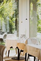Un comedor con mesas dispuestas para dos, ventanas amplias y lámparas colgantes.