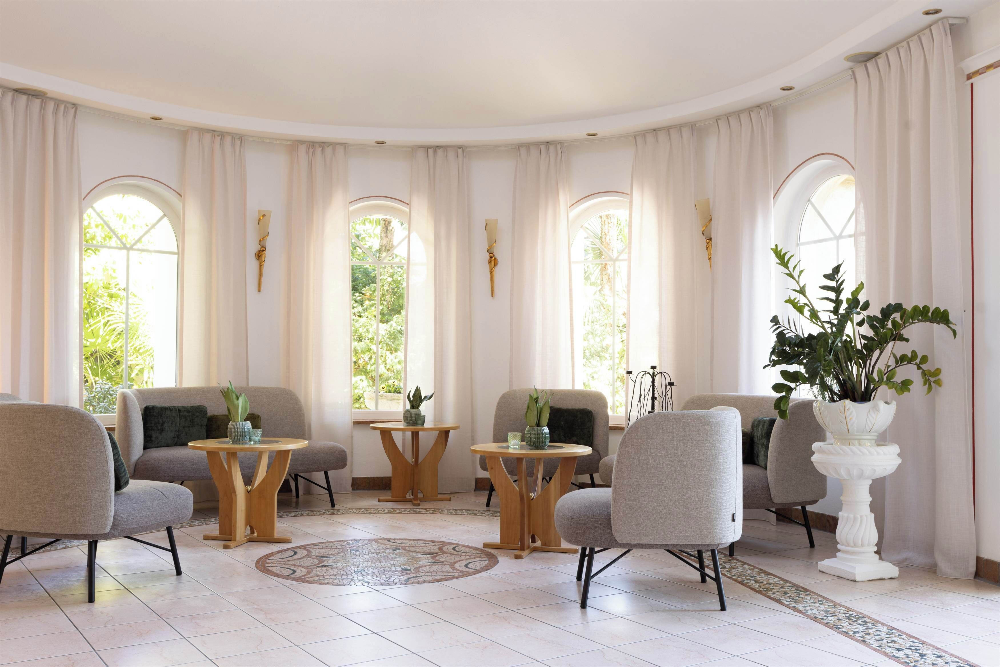 Una habitación amplia con techo abovedado, ventanas grandes con cortinas, varias zonas de estar y una planta decorativa como centro de atención.