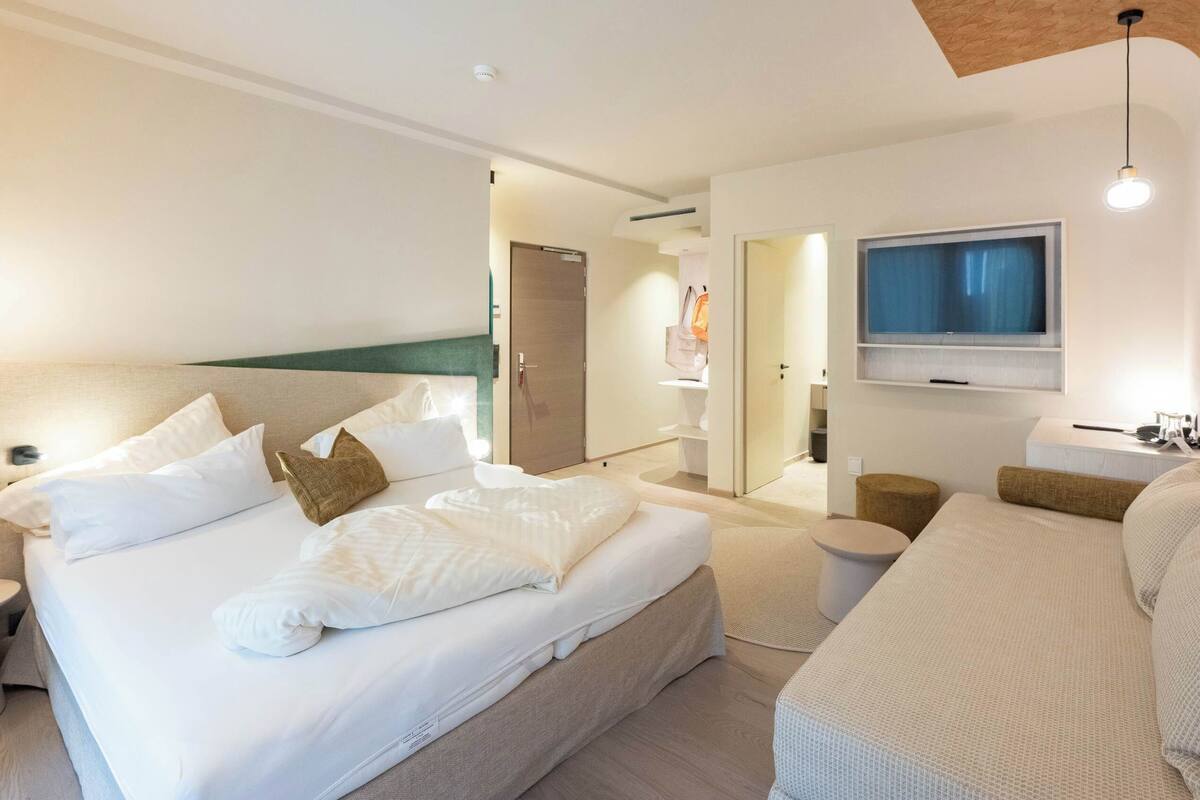 A modern hotel room with a large bed, bedside tables, a sofa, a TV, and a bathroom visible through an open door.