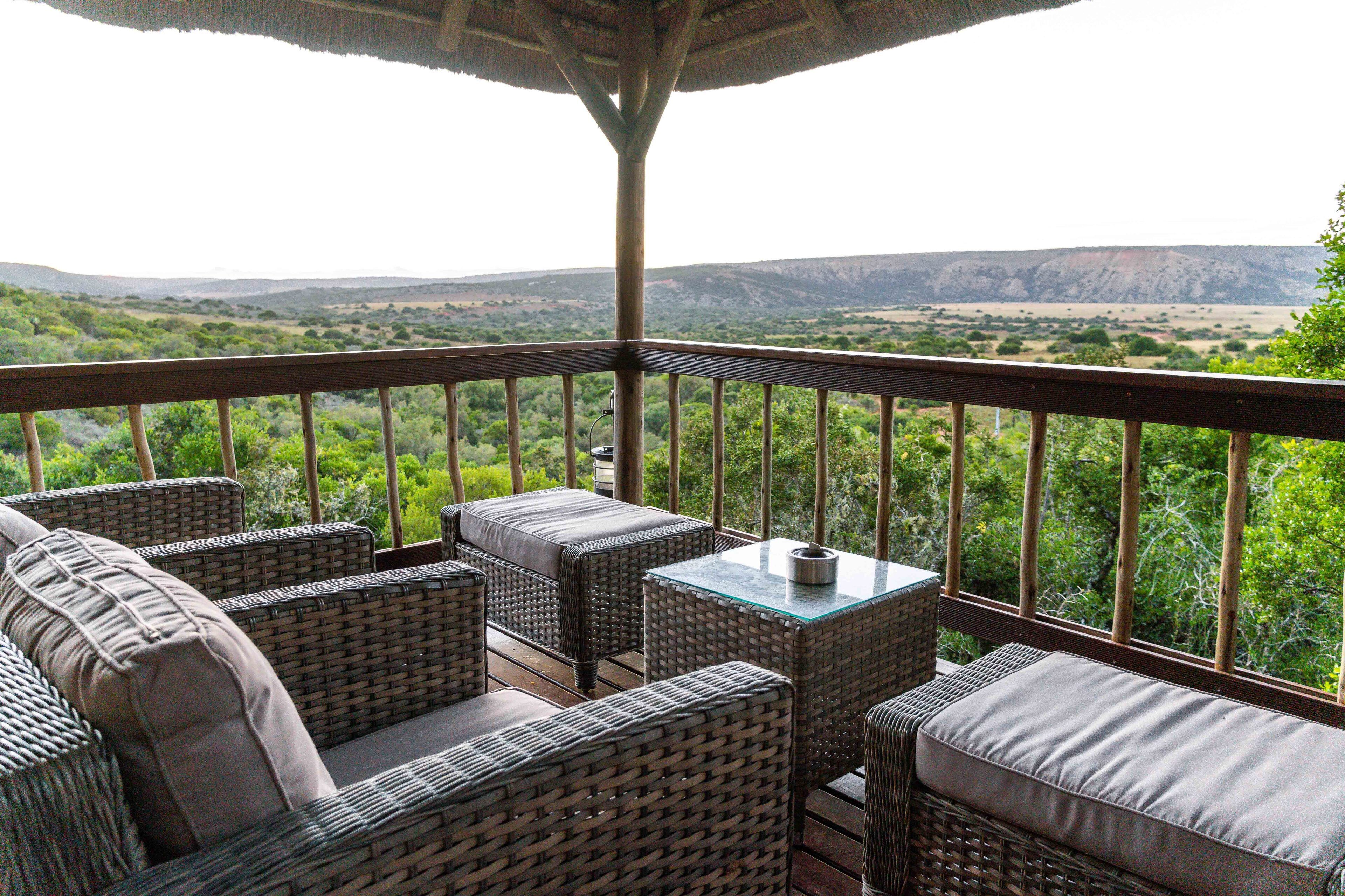 A covered outdoor seating area with wicker furniture and a glass table, overlooking a mountainous landscape.