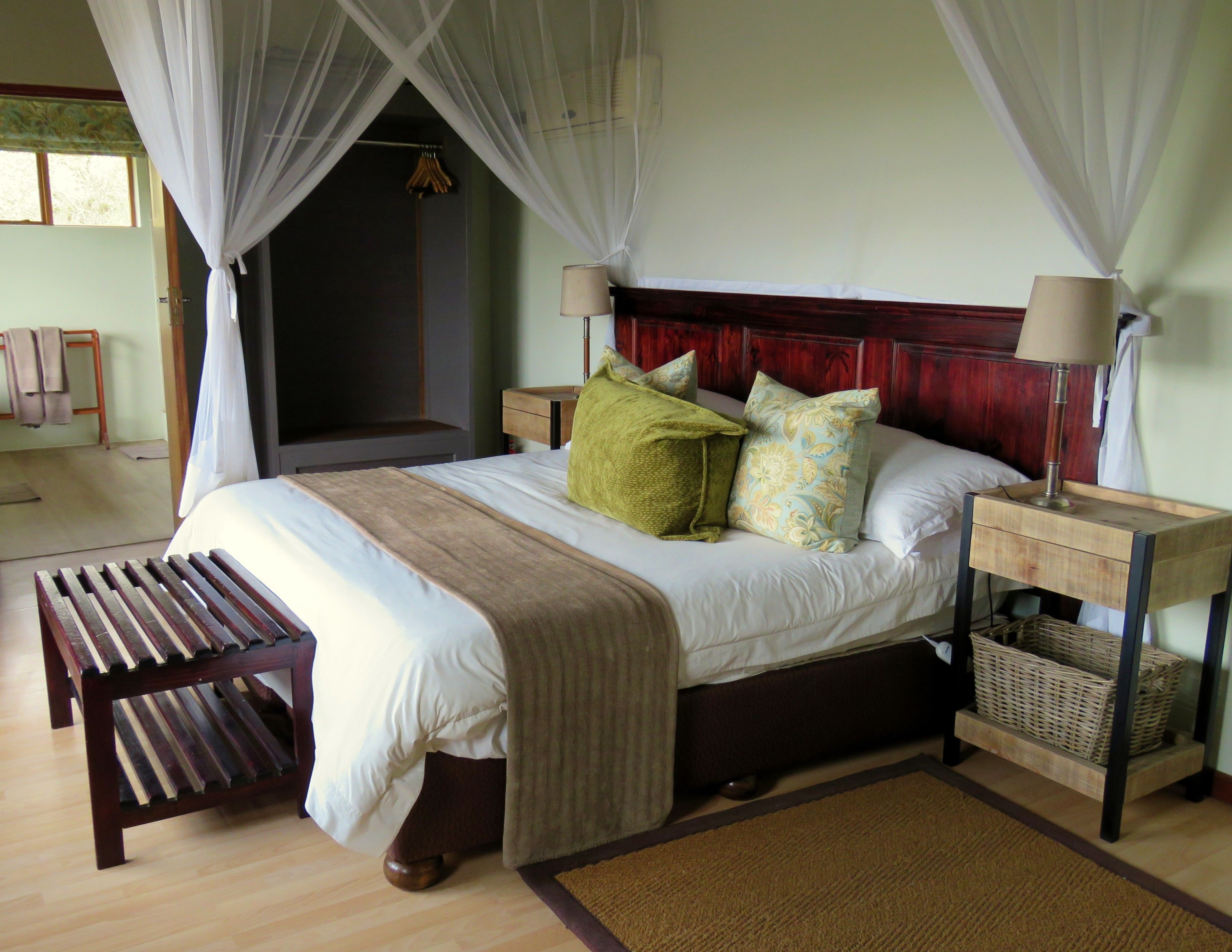 Un dormitorio con cama, mesitas de noche, un banco, una cesta junto a la cama y una ventana con cortinas.