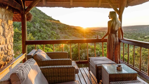 Una terraza de madera con muebles de mimbre y una mesa de vidrio, con vista a un paisaje exuberante al atardecer.