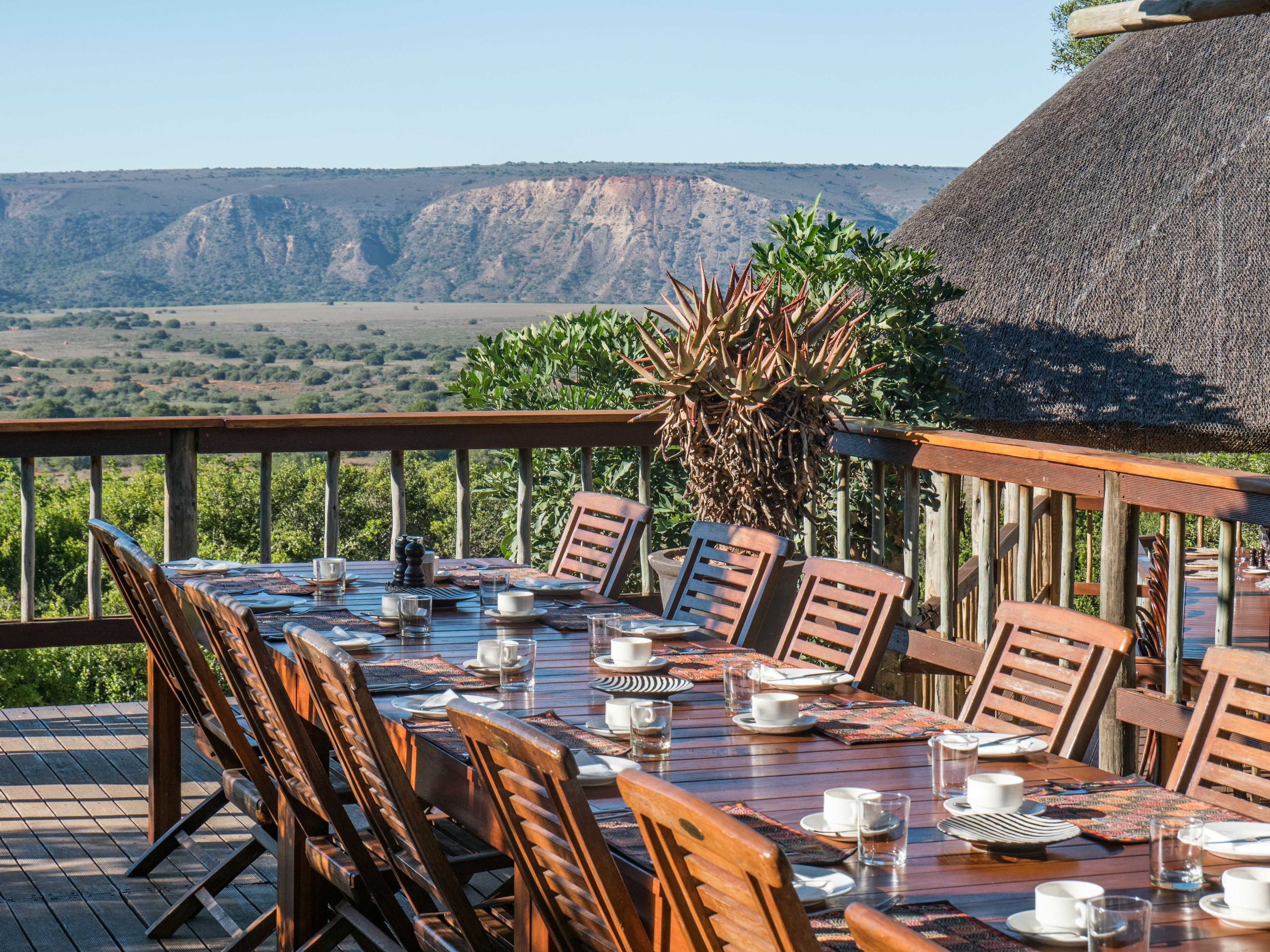 A dining table set outdoors with wooden chairs, a thatched roof, and a view of mountains in the background.