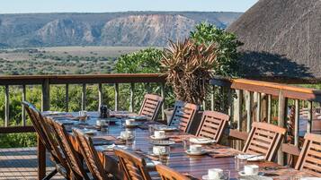 A dining table set outdoors with wooden chairs, a thatched roof, and a view of mountains in the background.