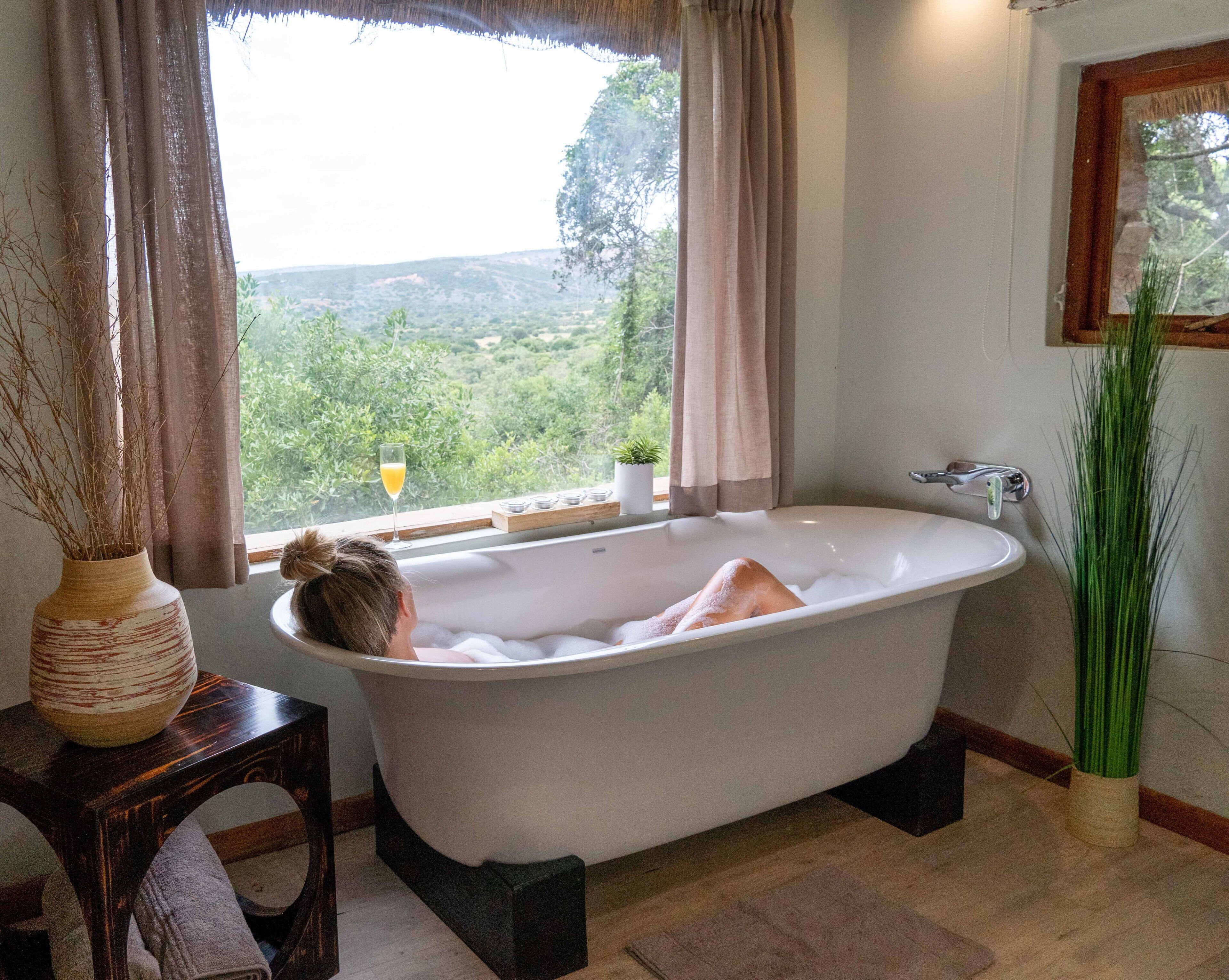 Un baño con una bañera independiente, una ventana con cortinas, un florero con ramas y una planta en maceta.