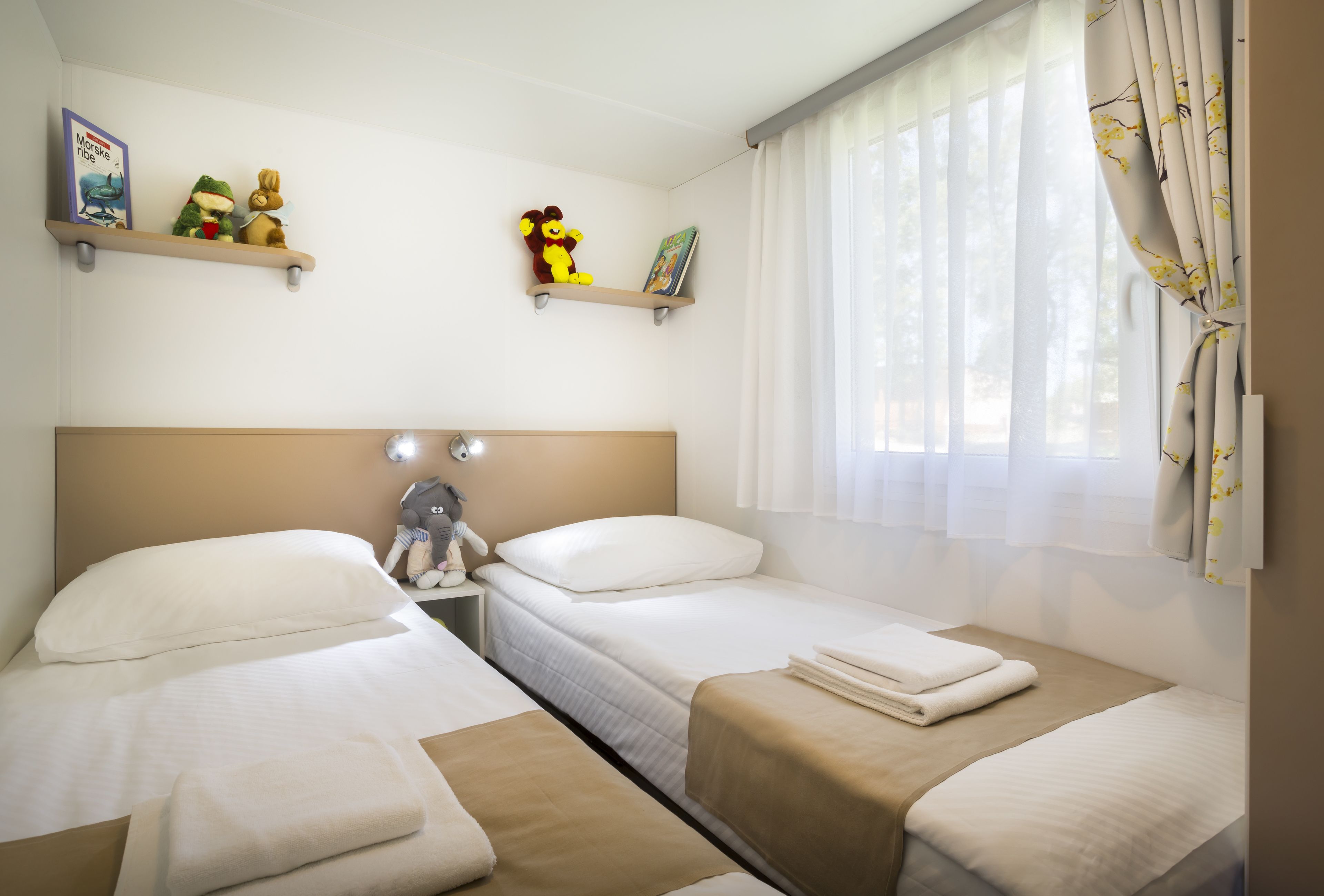 A hotel room with two single beds, a nightstand, a shelf with plush toys, and a window with sheer curtains.