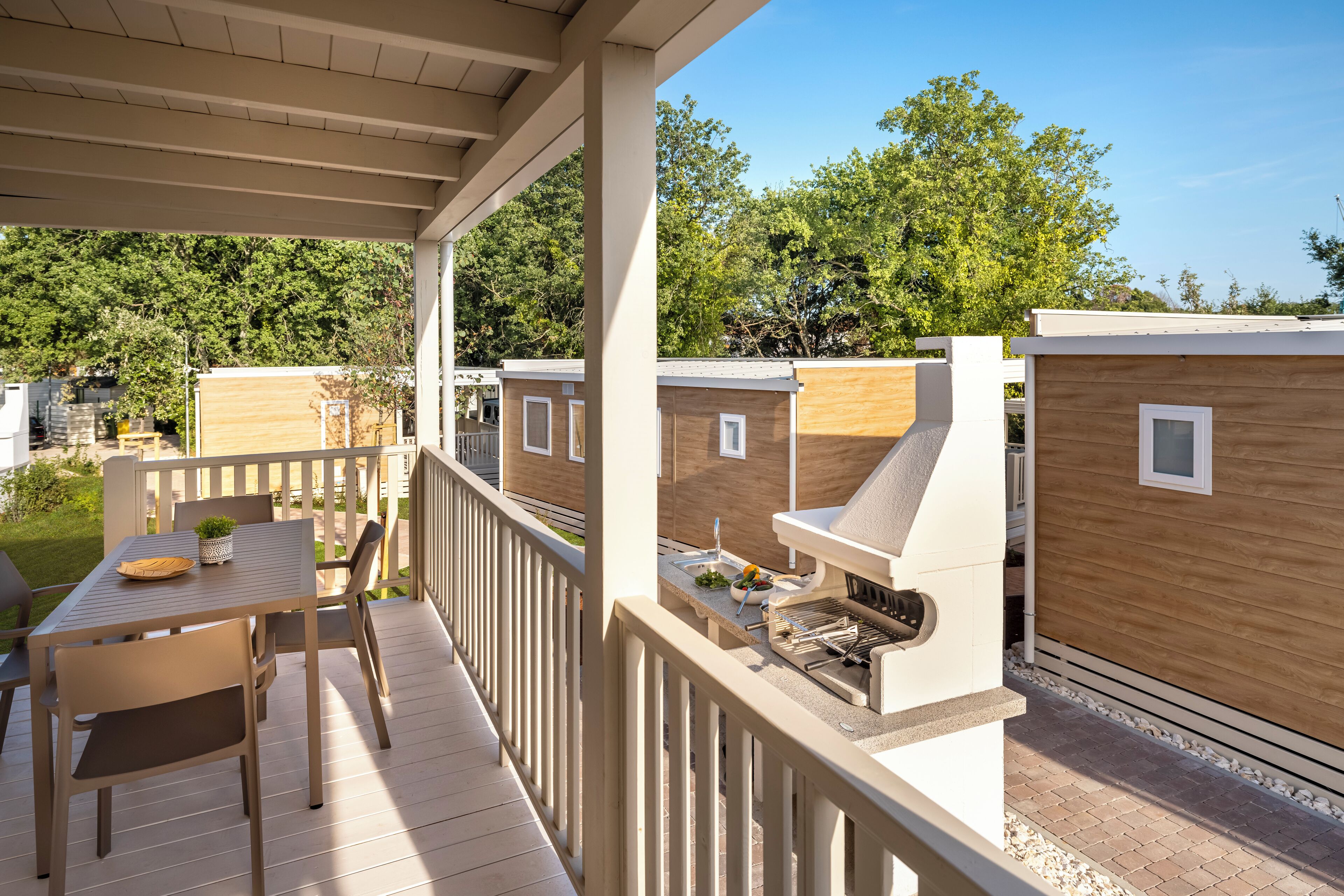 Eine überdachte Terrasse im Freien mit Tisch und Stühlen, einem Grill und Holzhütten im Hintergrund.