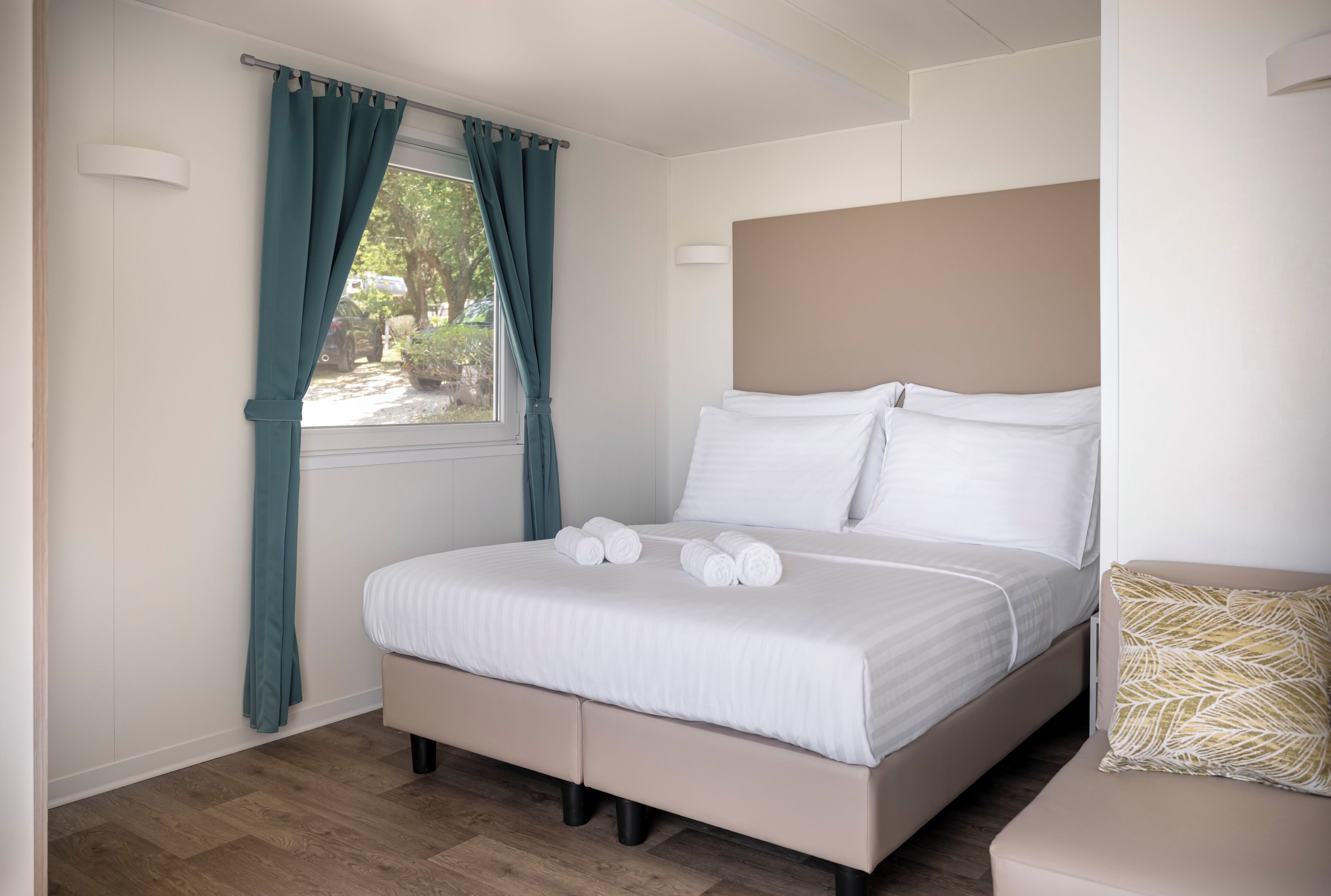 Una cama bien tendida con sábanas blancas y un cabecero, una ventana con cortinas color turquesa y un suelo de madera.