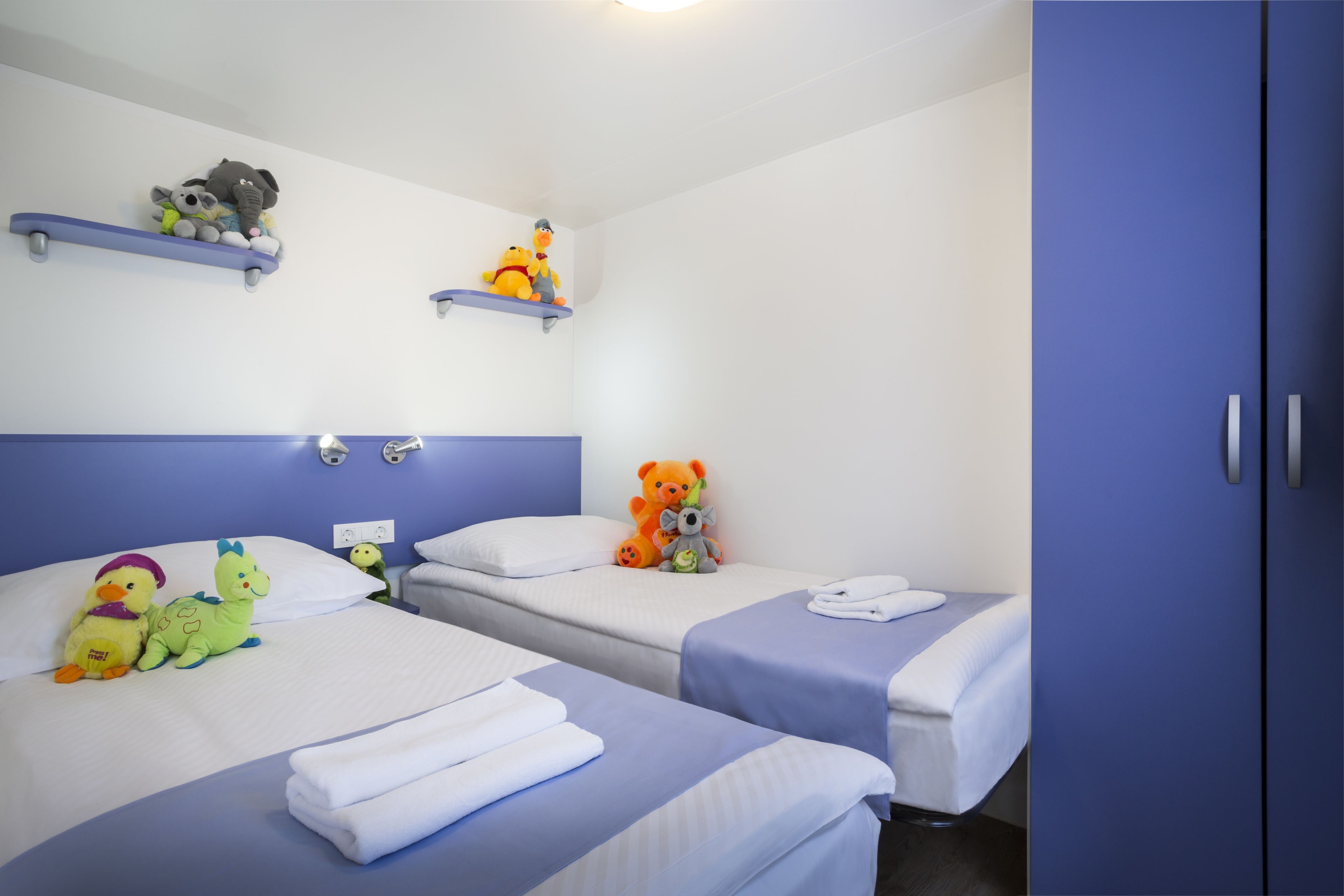 A hotel room with two beds, each with a blue bedspread and white pillows. There are plush toys on the beds and shelves above the beds.