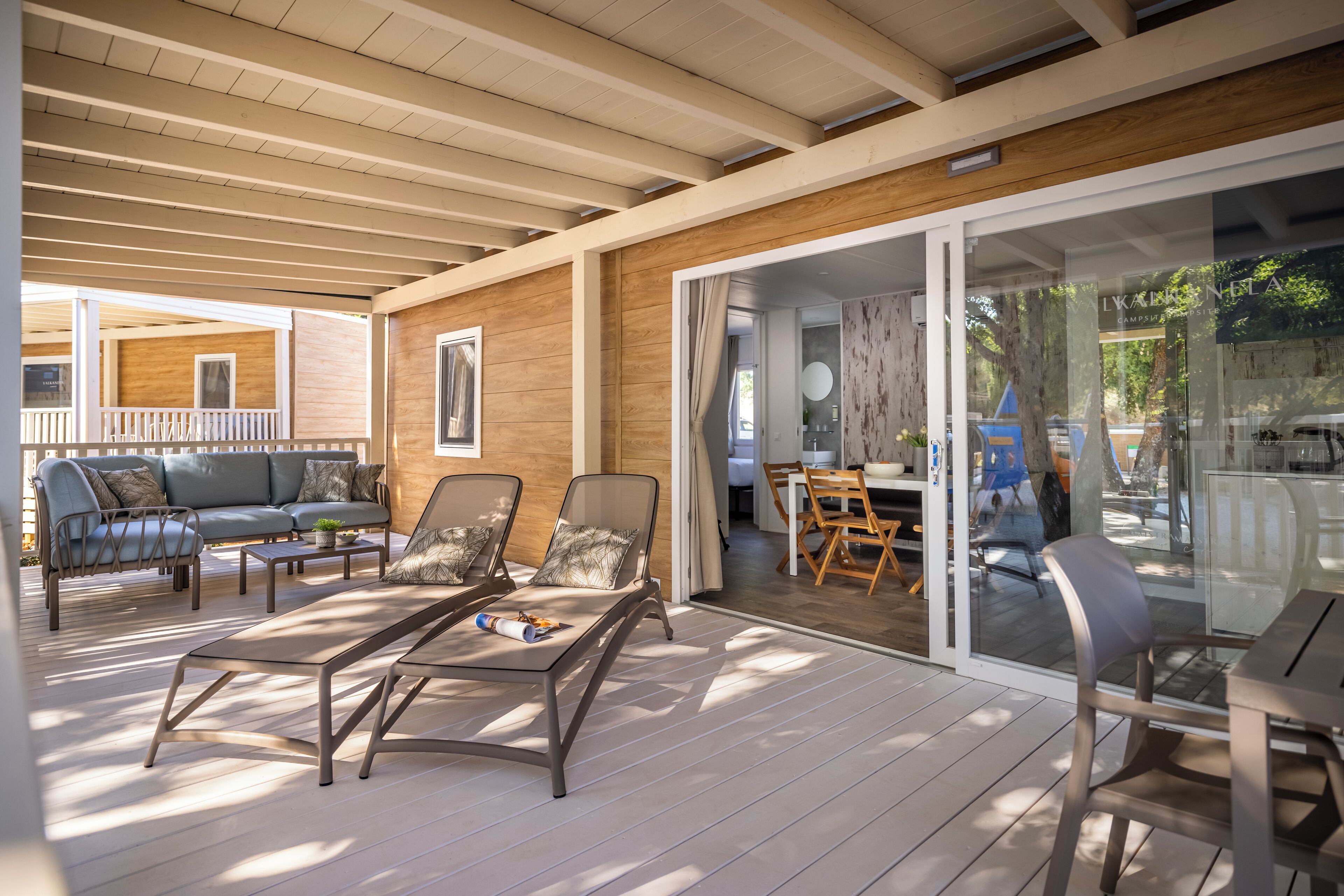 A covered outdoor area with a wooden deck, a seating area with a sofa and chairs, and a glass door leading to a dining area.