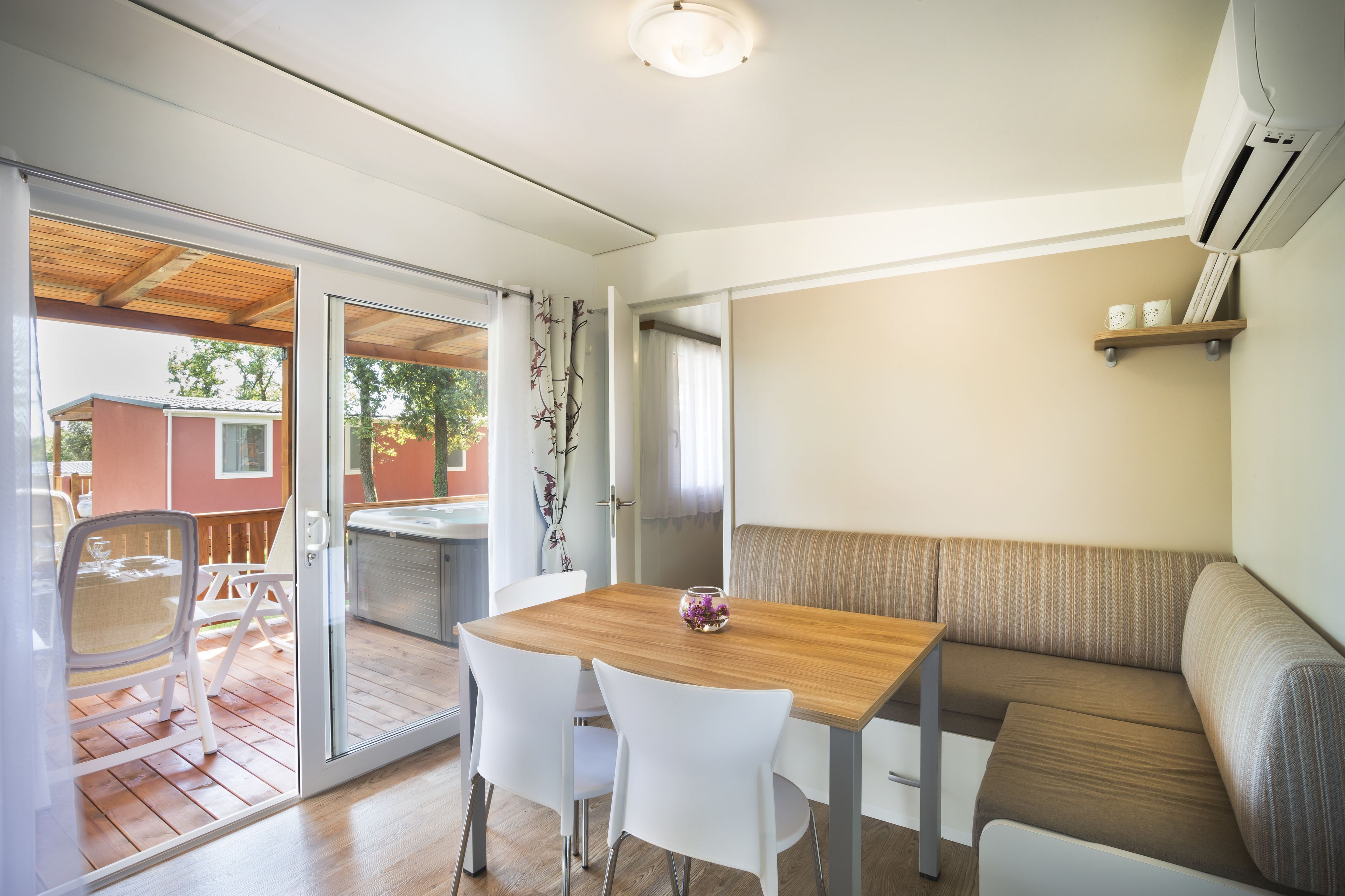 A modern room with a wooden table, white chairs, a cushioned bench, and a sliding glass door leading to an outdoor area with a patio and a view of a neighboring house.