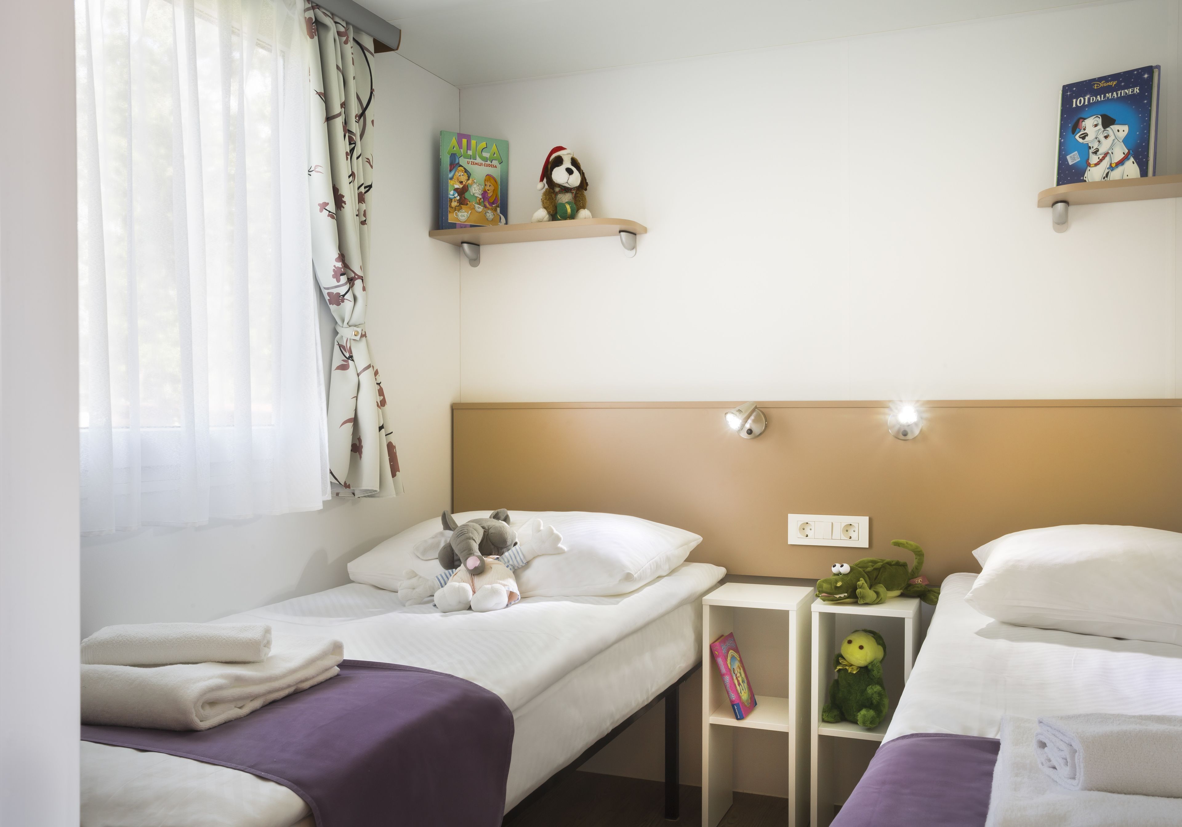 Een hotelkamer met twee enkele bedden, een raam met dunne gordijnen, een plank met boeken en een pluche knuffel, en een wandlamp.