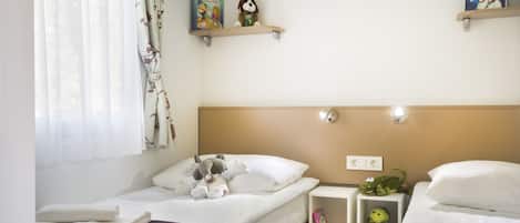 A hotel room with two single beds, a window with sheer curtains, a shelf with books and a stuffed animal, and a wall-mounted light fixture.