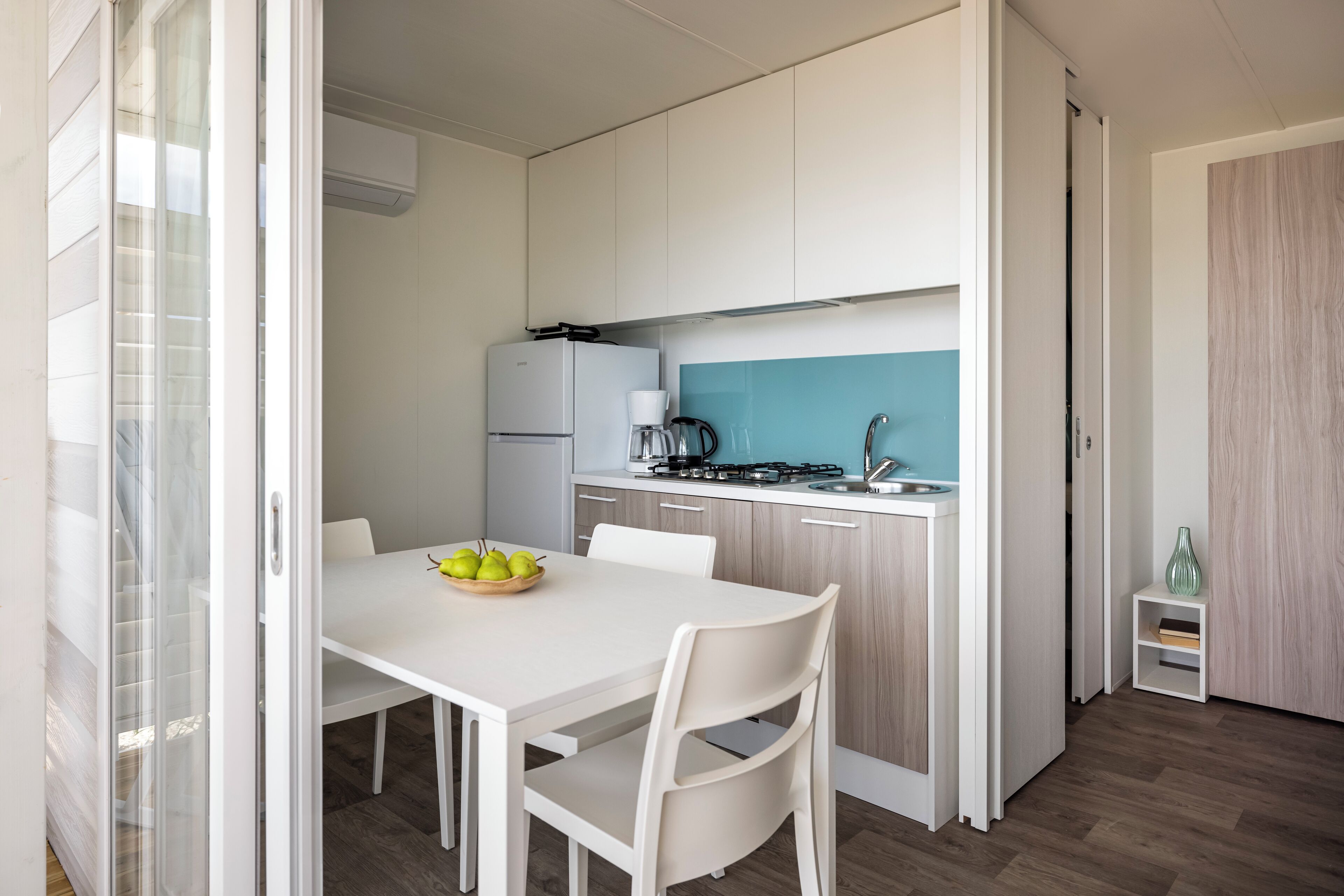 Een moderne keuken met witte kasten, een wit aanrecht en een gasfornuis. Er staat een witte eettafel met stoelen en een schaal met fruit.