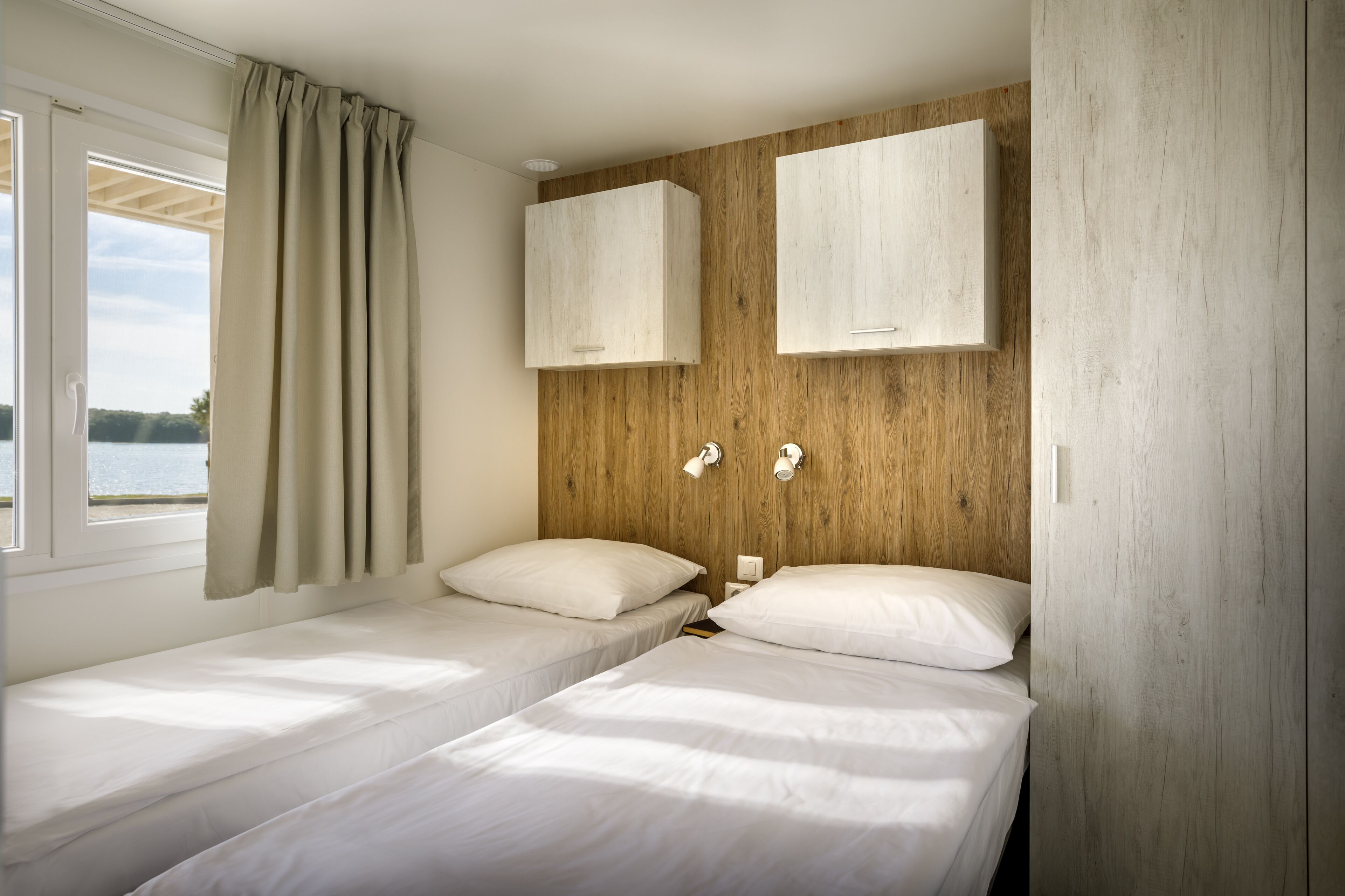 Habitació amb dos llits individuals, fusta al capçal, parets blanques i una finestra amb cortines.