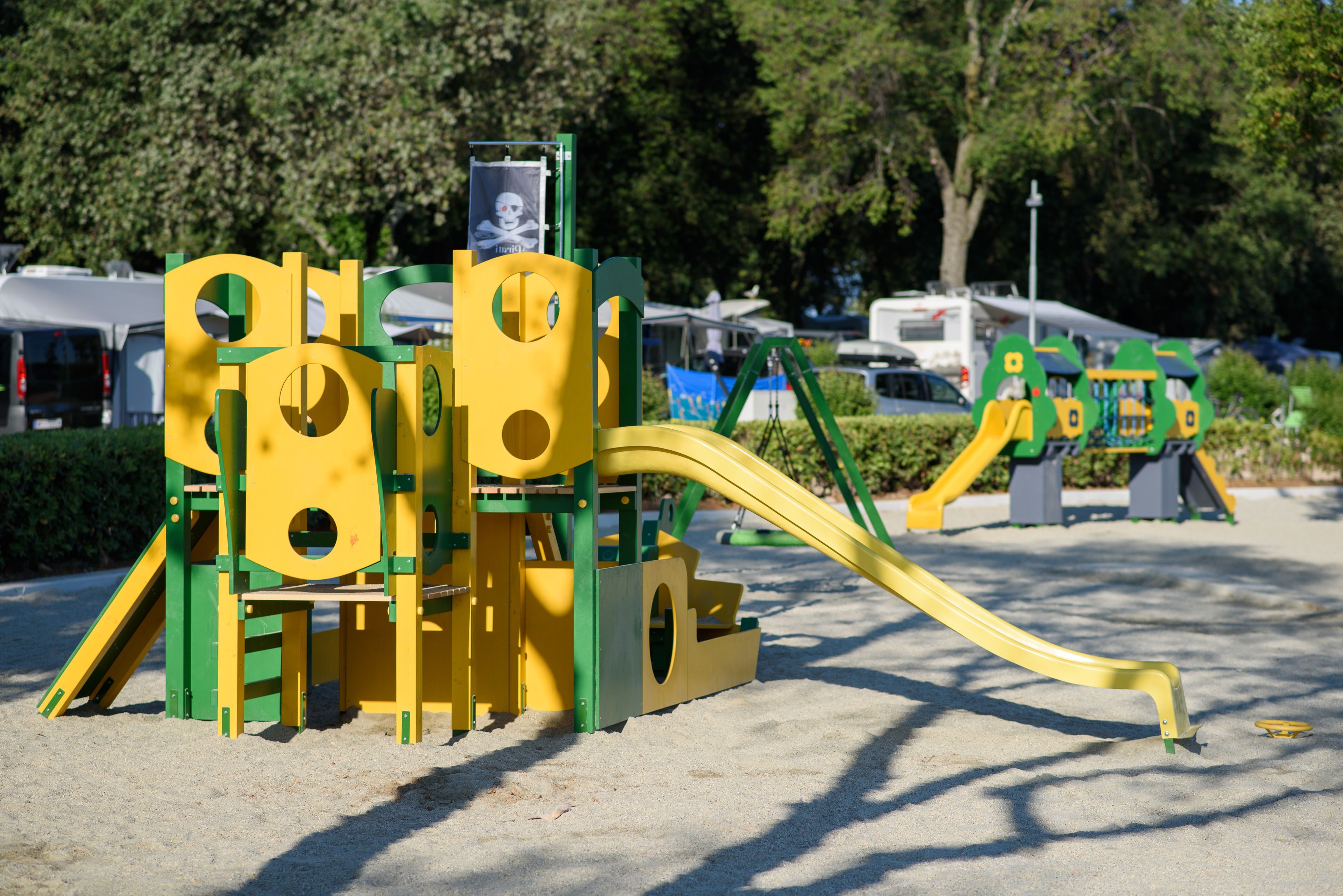 Un terrain de jeux avec des structures colorées en jaune et en vert, comprenant des toboggans et des zones d’escalade, aménagé dans un espace sablonneux avec des arbres en arrière-plan.