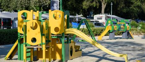 A playground with yellow and green play structures, including slides and climbing areas, set in a sandy area with trees in the background.