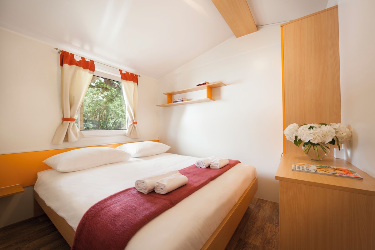 A neatly made bed with white linens and a red blanket, a wooden nightstand with a vase of white flowers, and a window with orange curtains.