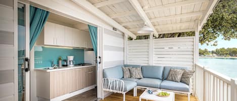 A modern outdoor living area with a wooden deck, a white sofa with blue cushions, a white coffee table, and a kitchen area with a teal backsplash.