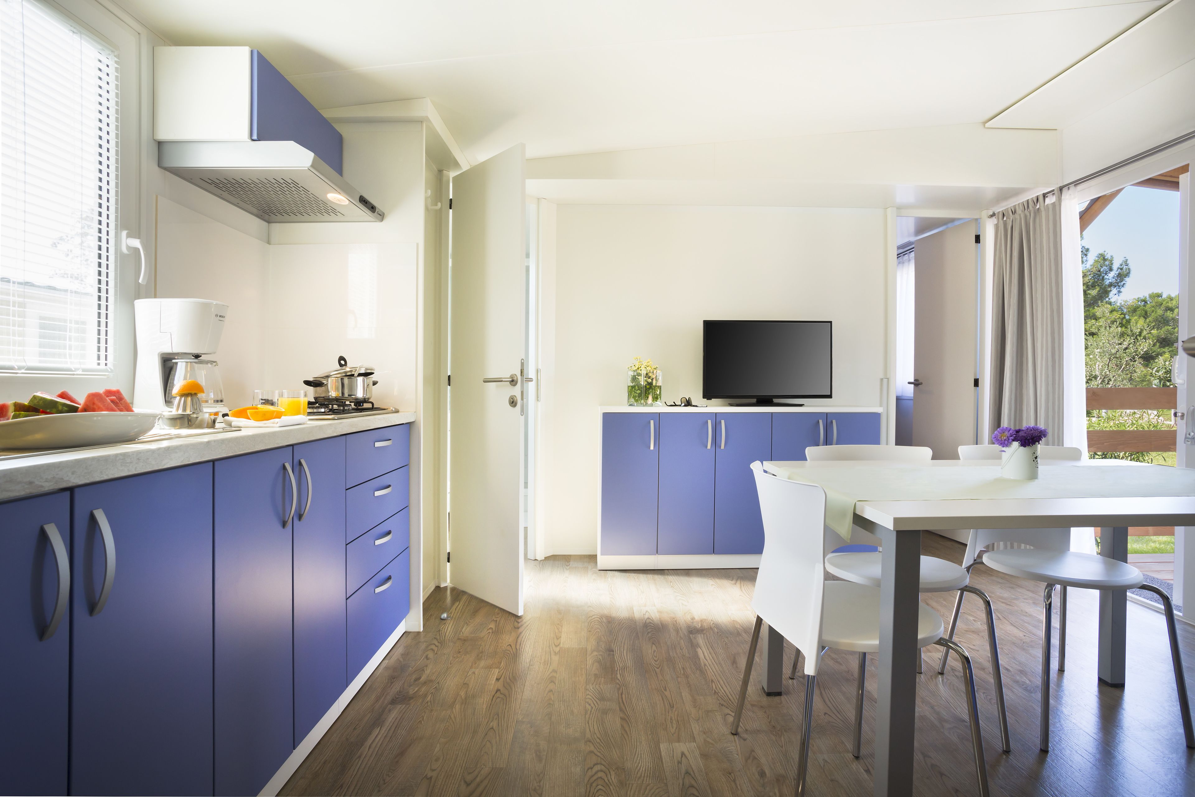 A modern kitchen with blue cabinets, a white dining table, and a TV in the background.