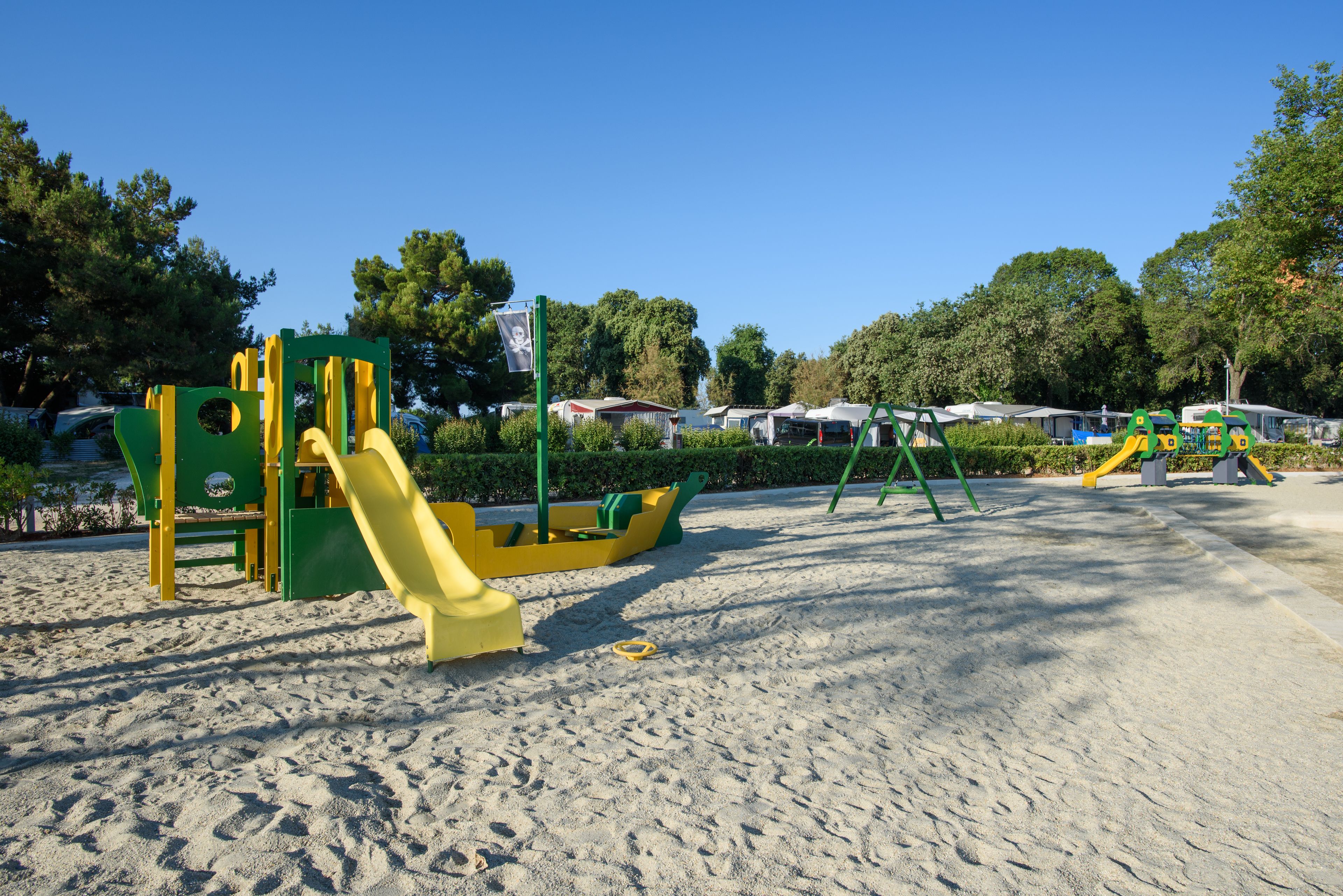 Un terrain de jeux avec du sable, des structures colorées en vert et en jaune, et des arbres en arrière-plan.