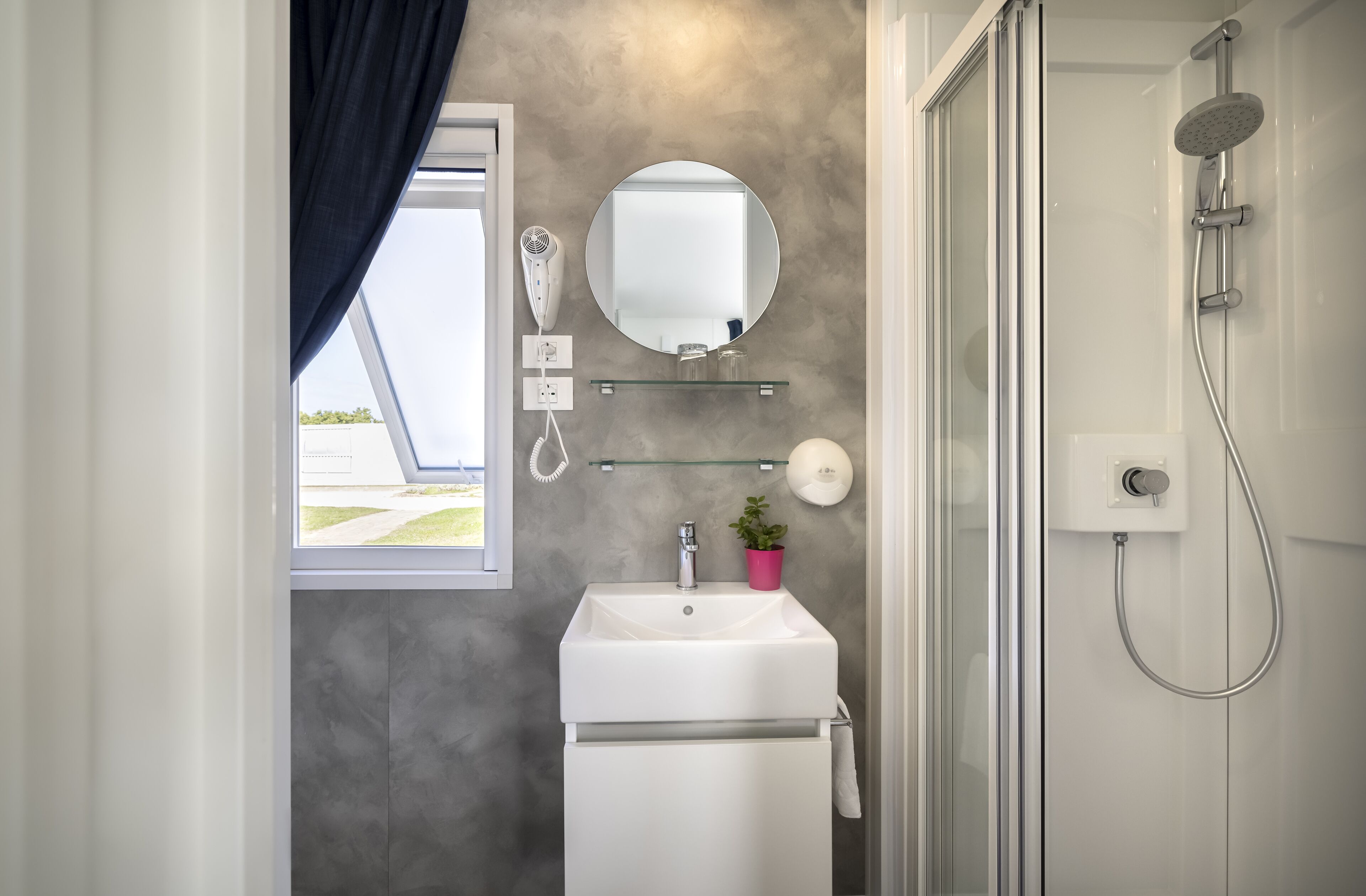 A bathroom with a white sink, a round mirror, a glass shelf with toiletries, a shower with a handheld showerhead, and a window with a curtain.