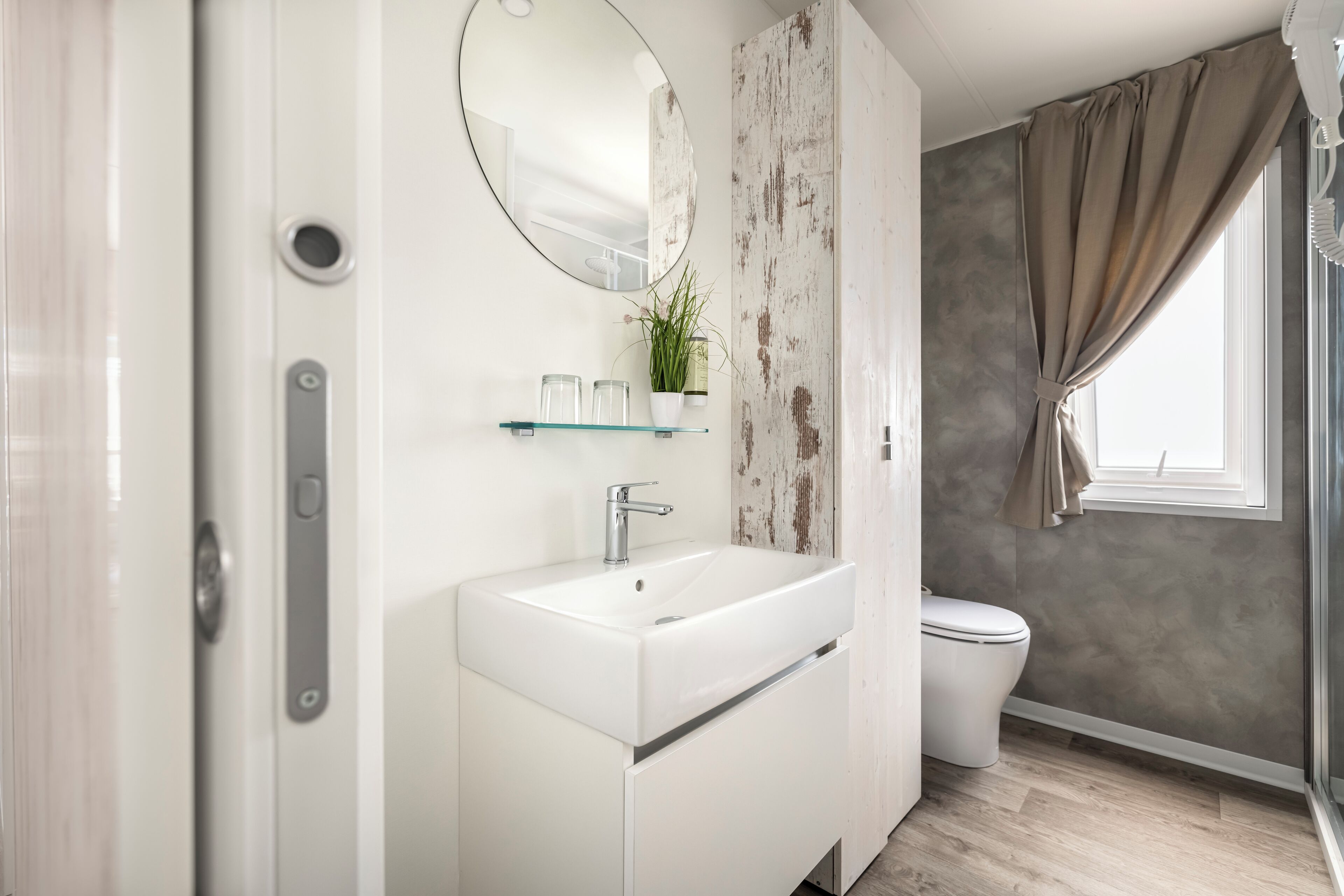 A modern bathroom with a white vanity, a round mirror, and a glass shelf.