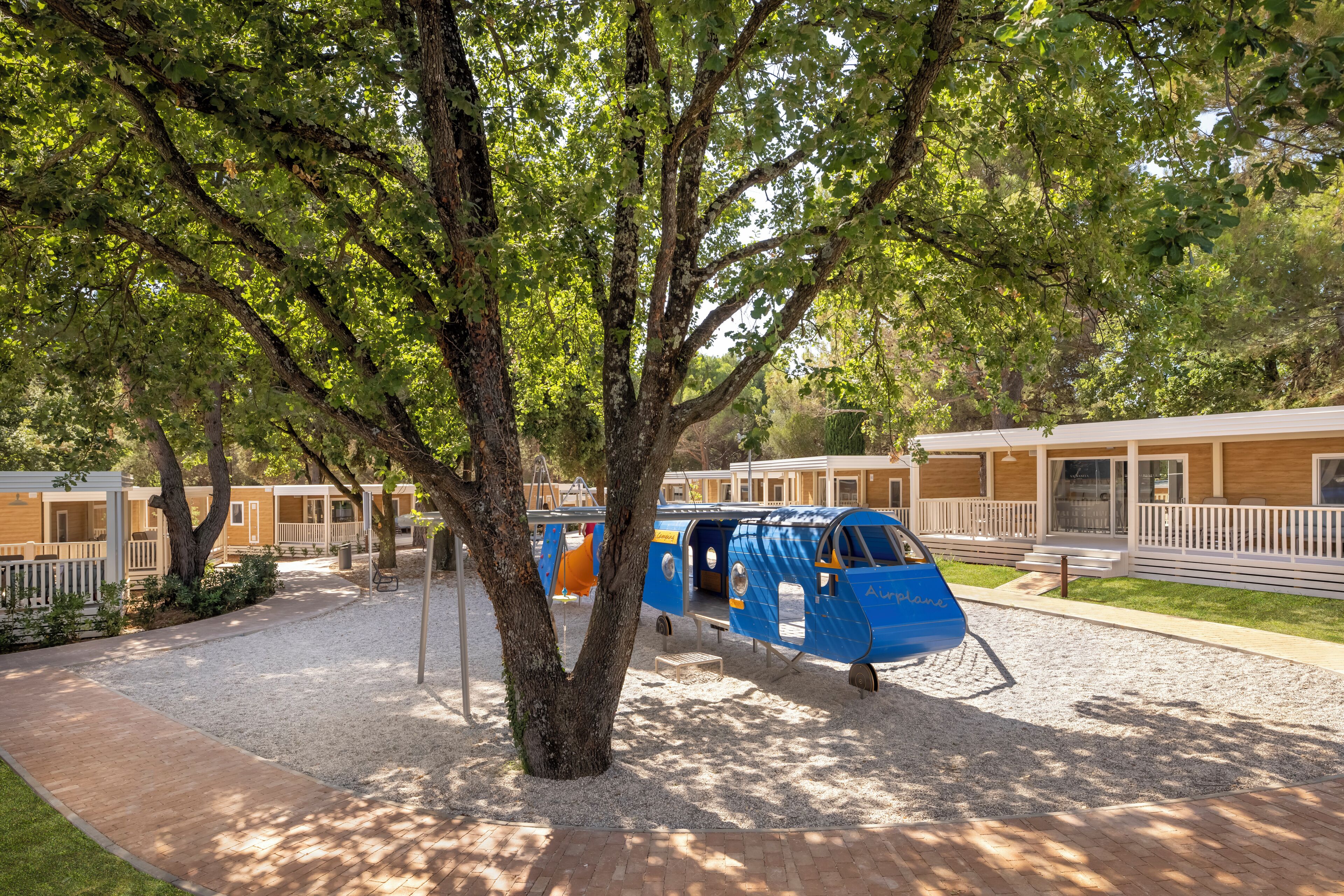 Un avión azul estacionado bajo un árbol en un camping con cabañas al fondo.
