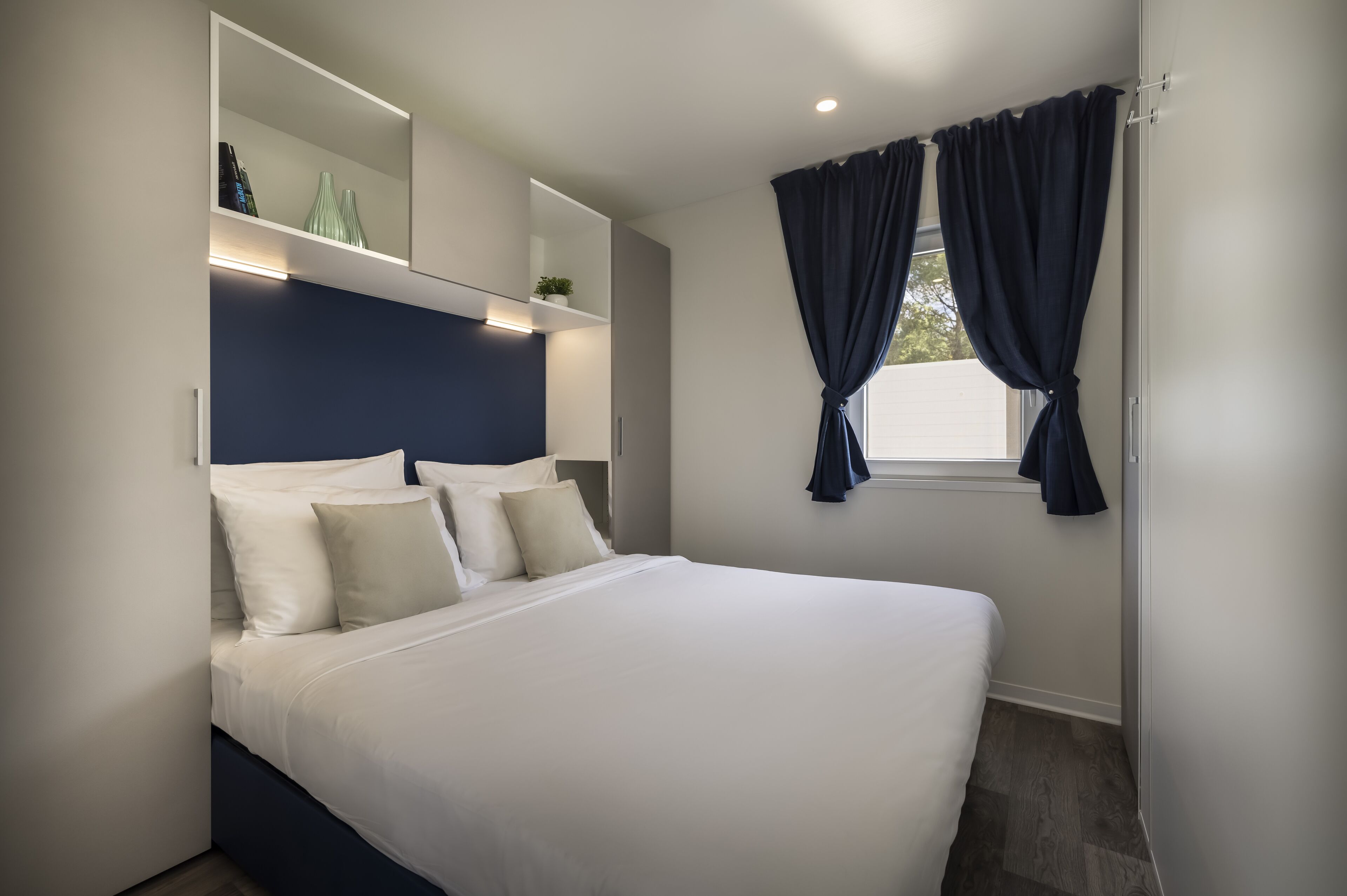 Un dormitorio moderno con una cama grande, una mesita de noche empotrada, un espejo y una ventana con cortinas.