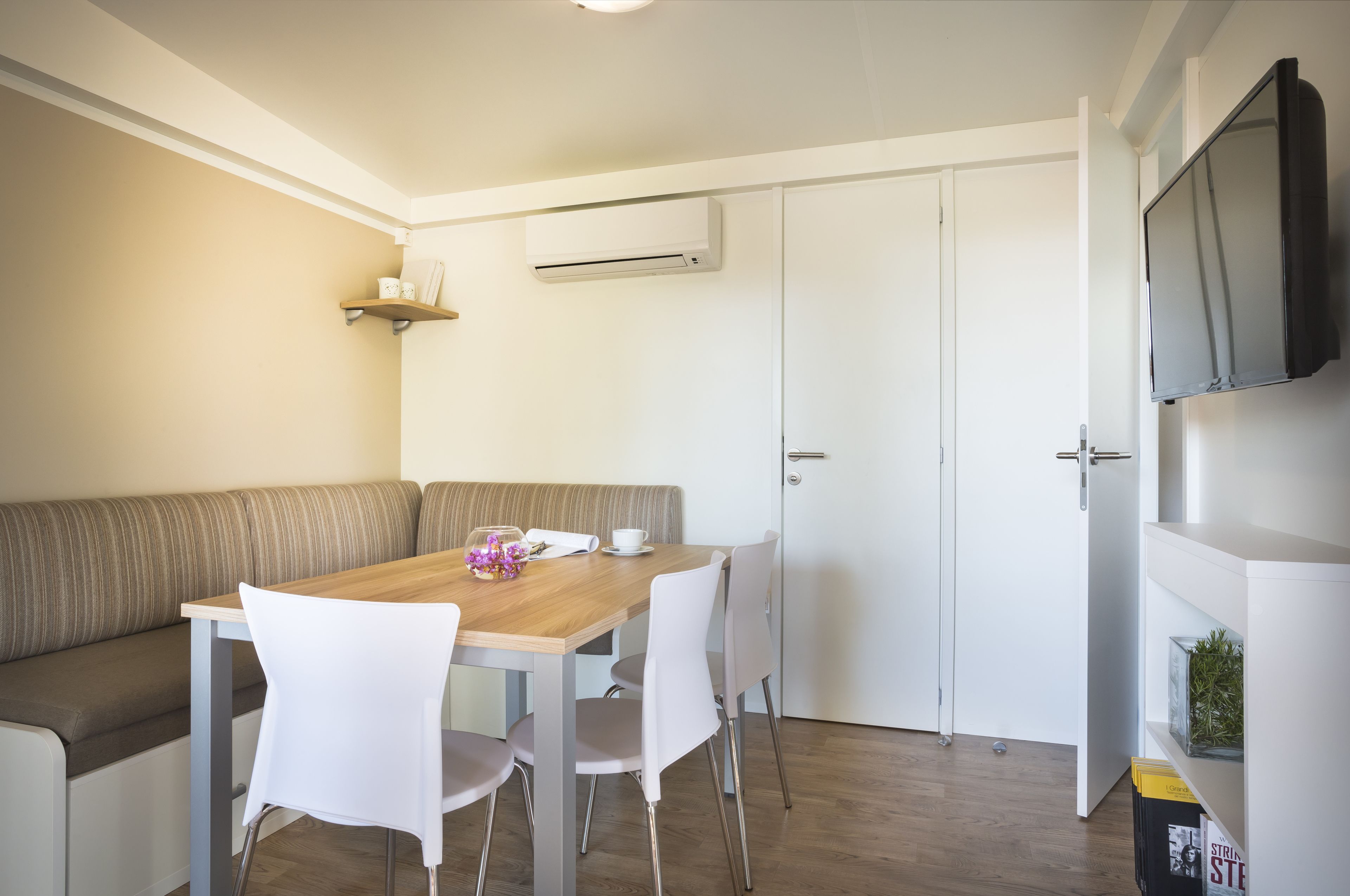 Een moderne keuken met een houten tafel, witte stoelen, een inbouwbank en een aan de muur bevestigde televisie.