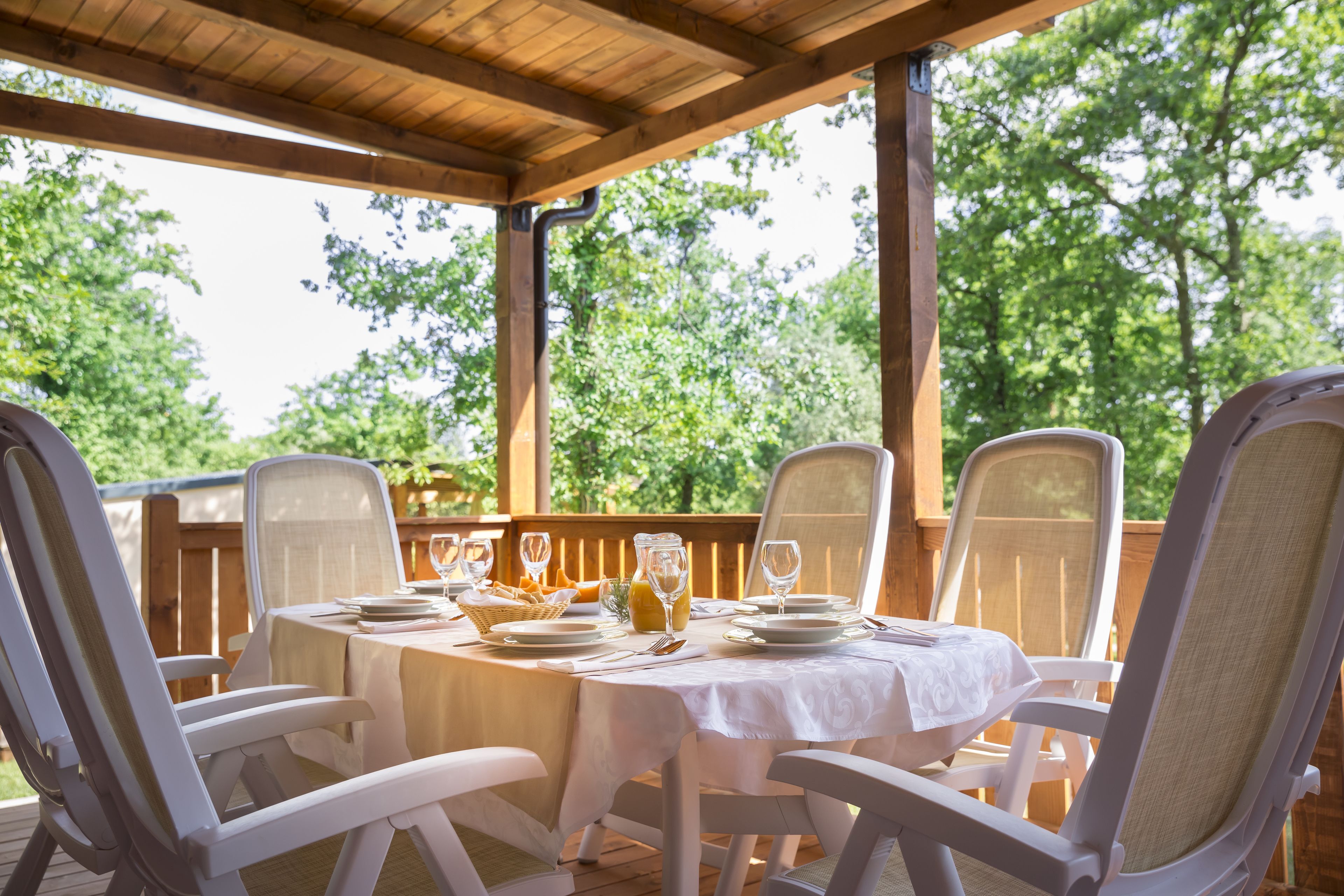 Un espacio de comedor al aire libre techado, con una mesa dispuesta para cuatro personas, rodeada de sillas blancas y una terraza de madera con vista a árboles.