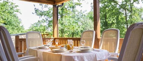 Un espacio de comedor al aire libre techado, con una mesa dispuesta para cuatro personas, rodeada de sillas blancas y una terraza de madera con vista a árboles.