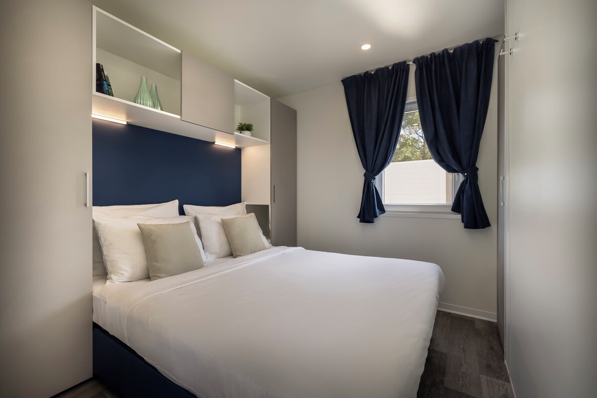 A modern bedroom with a large bed, a built-in nightstand, a mirror, and a window with curtains.