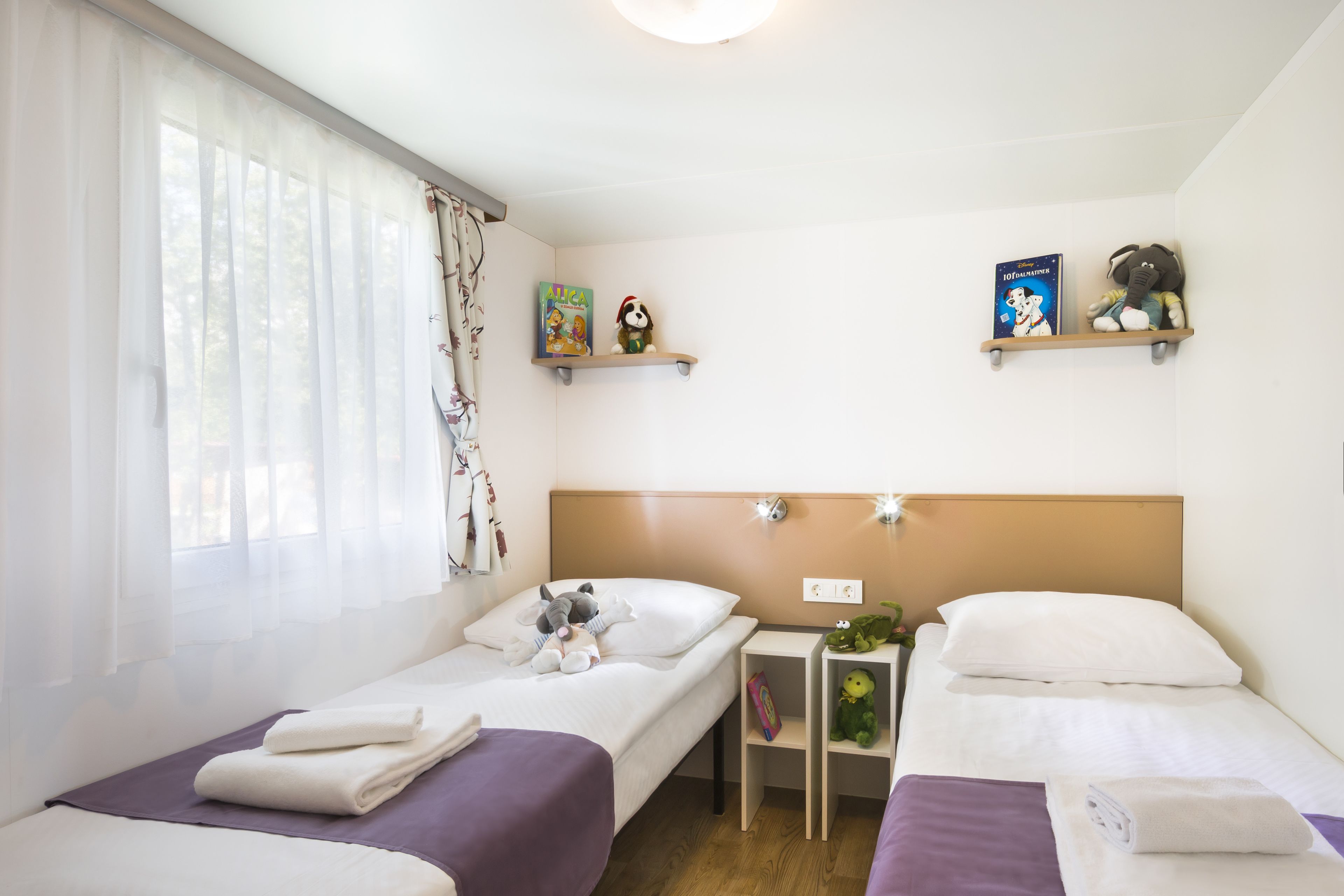 Una camera d'albergo con due letti, una finestra con tende leggere, un tavolino con una pianta e due mensole con libri e peluche.