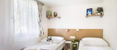 Habitación de hotel con dos camas, una ventana con cortinas translúcidas, una mesita con una planta y dos estantes con libros y peluches.