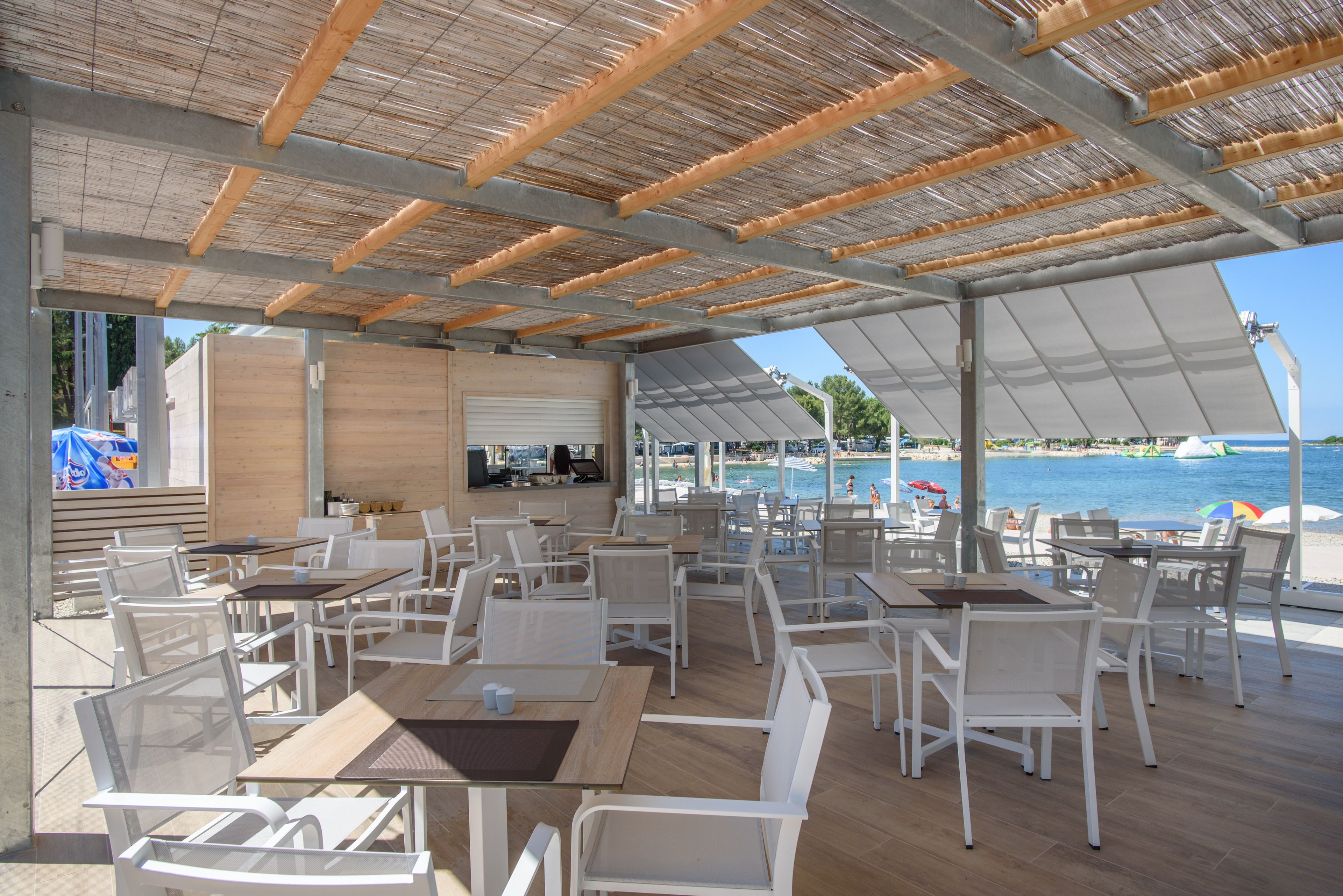 Un espace extérieur aménagé pour les repas, avec des chaises et des tables blanches, une terrasse en bois et une vue sur la mer.