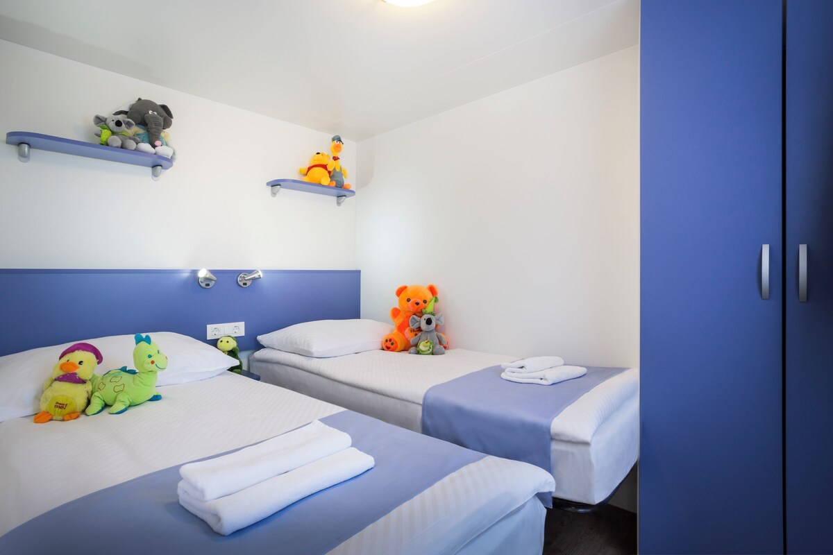 A hotel room with two beds, each with a blue bedspread and white pillows. There are plush toys on the beds and shelves above the beds.