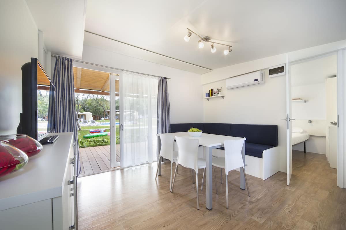 Un salon moderne et bien éclairé, comprenant une table à manger, des chaises blanches, un canapé de couleur foncée et une porte vitrée donnant sur un espace extérieur.