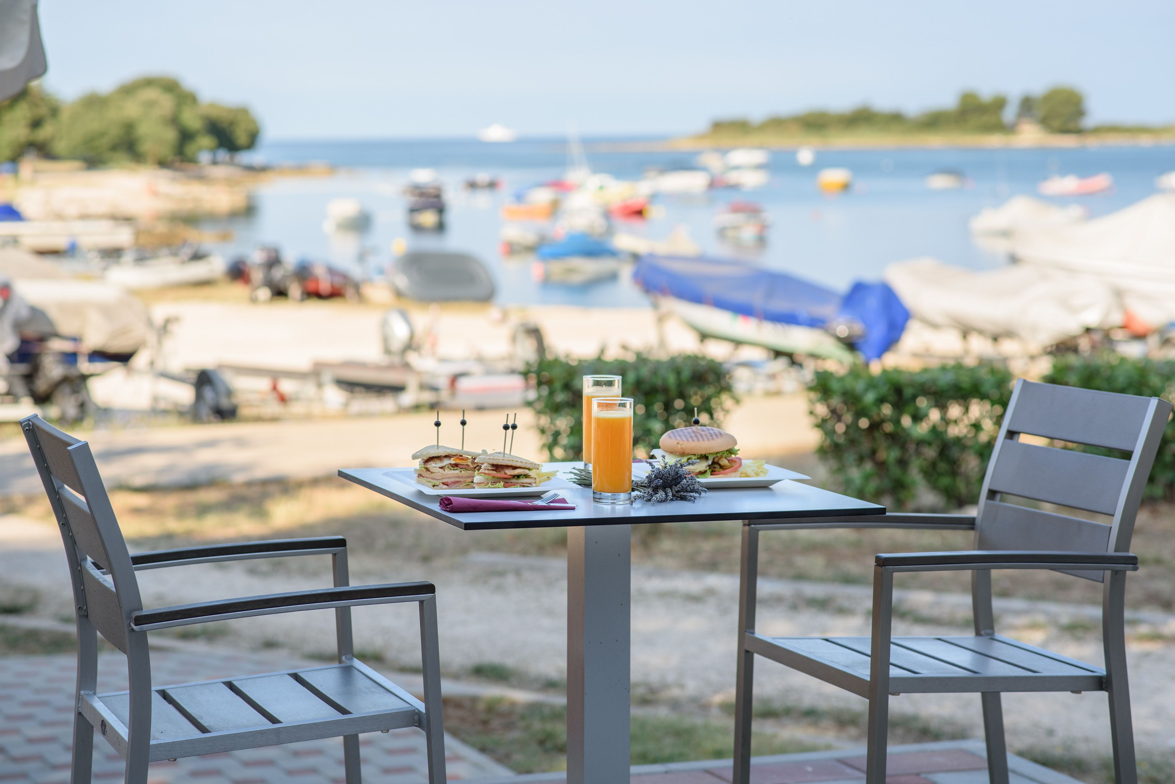 Ett bord med två stolar, en smörgås, en hamburgare och ett glas apelsinjuice, med utsikt över en strand med båtar och parasoller.