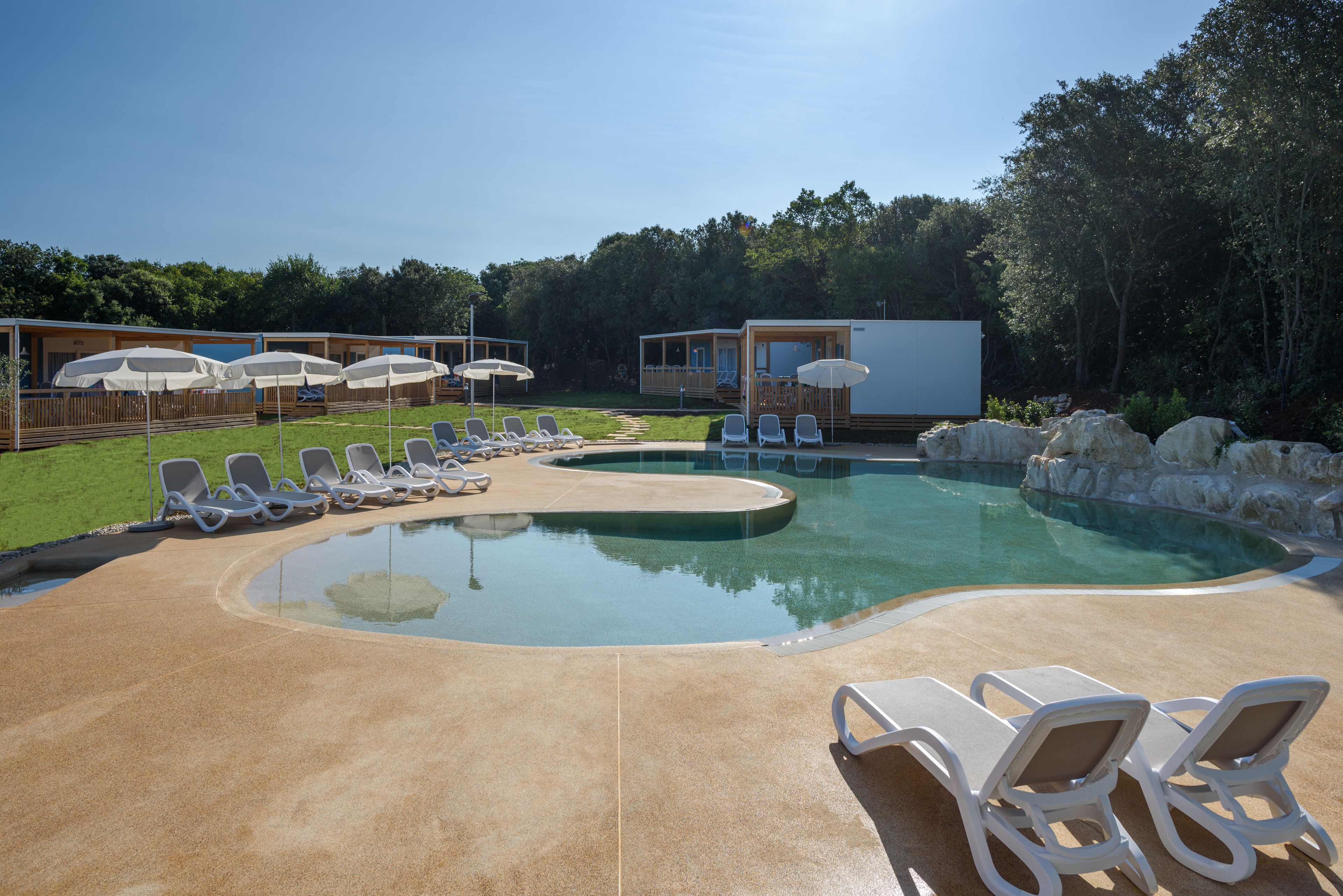 Una piscina con sillones de descanso y sombrillas, rodeada por una terraza de madera y una zona ajardinada.
