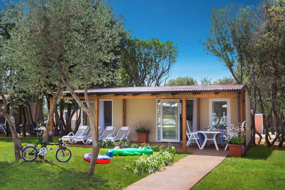 Un petit bâtiment d’un seul étage, doté d’un toit en tuiles, entouré d’arbres et d’une pelouse verdoyante. On y trouve plusieurs chaises longues et un vélo garé à l’extérieur.