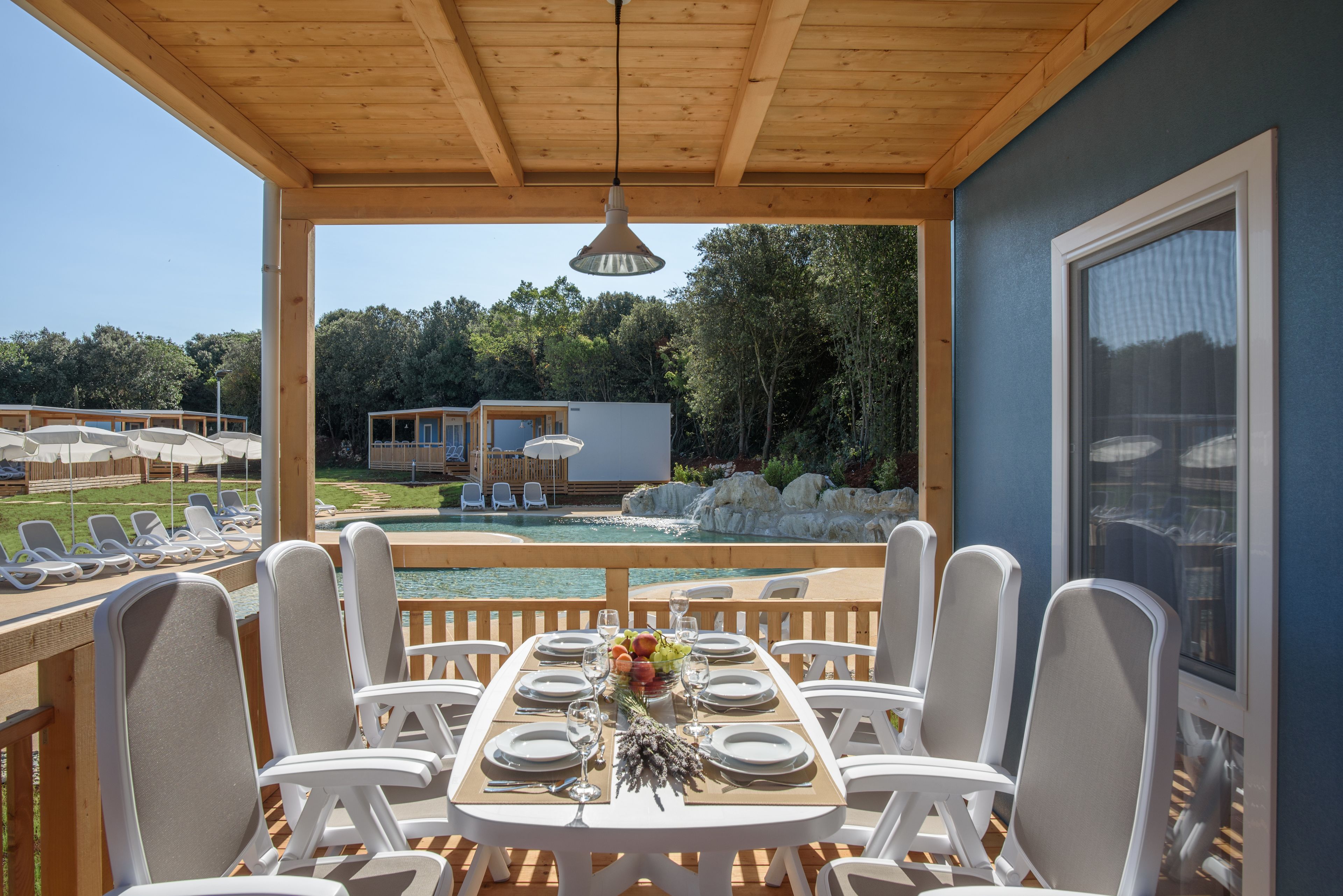 A covered outdoor dining area with a table set for six, overlooking a pool and a forested area.